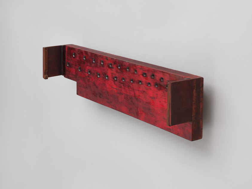 A long, rectangular red wooden object with two end panels and rows of black knobs mounted on a wall.