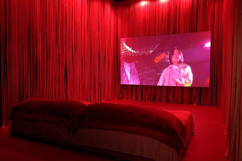 A red-lit room with a bed and a large screen showing a person in a white shirt.