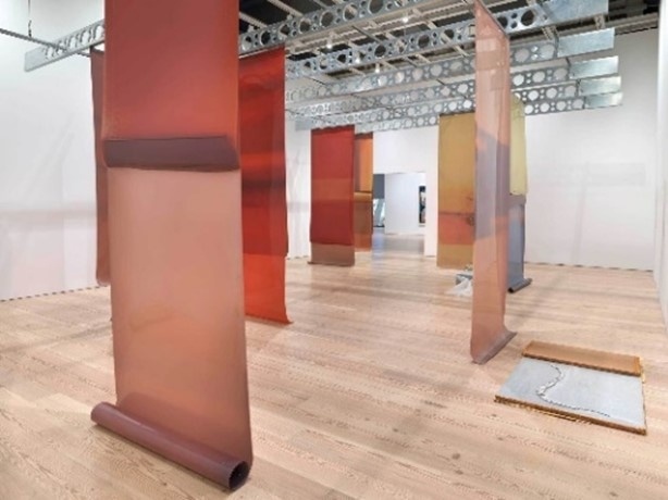 Large translucent colored panels hang vertically in a gallery space with wooden floors and metal ceiling beams.