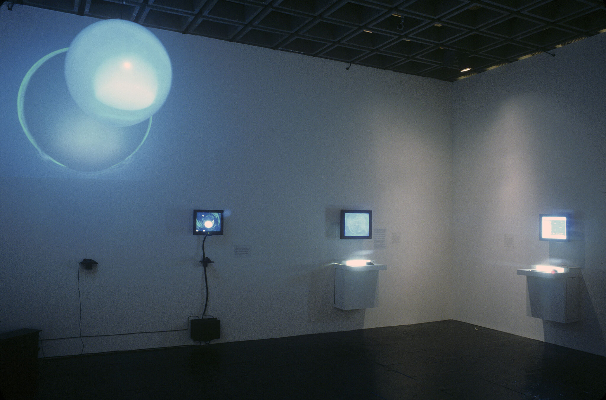 A gallery with a blue-hued video projection, along with monitors mounted on the walls.