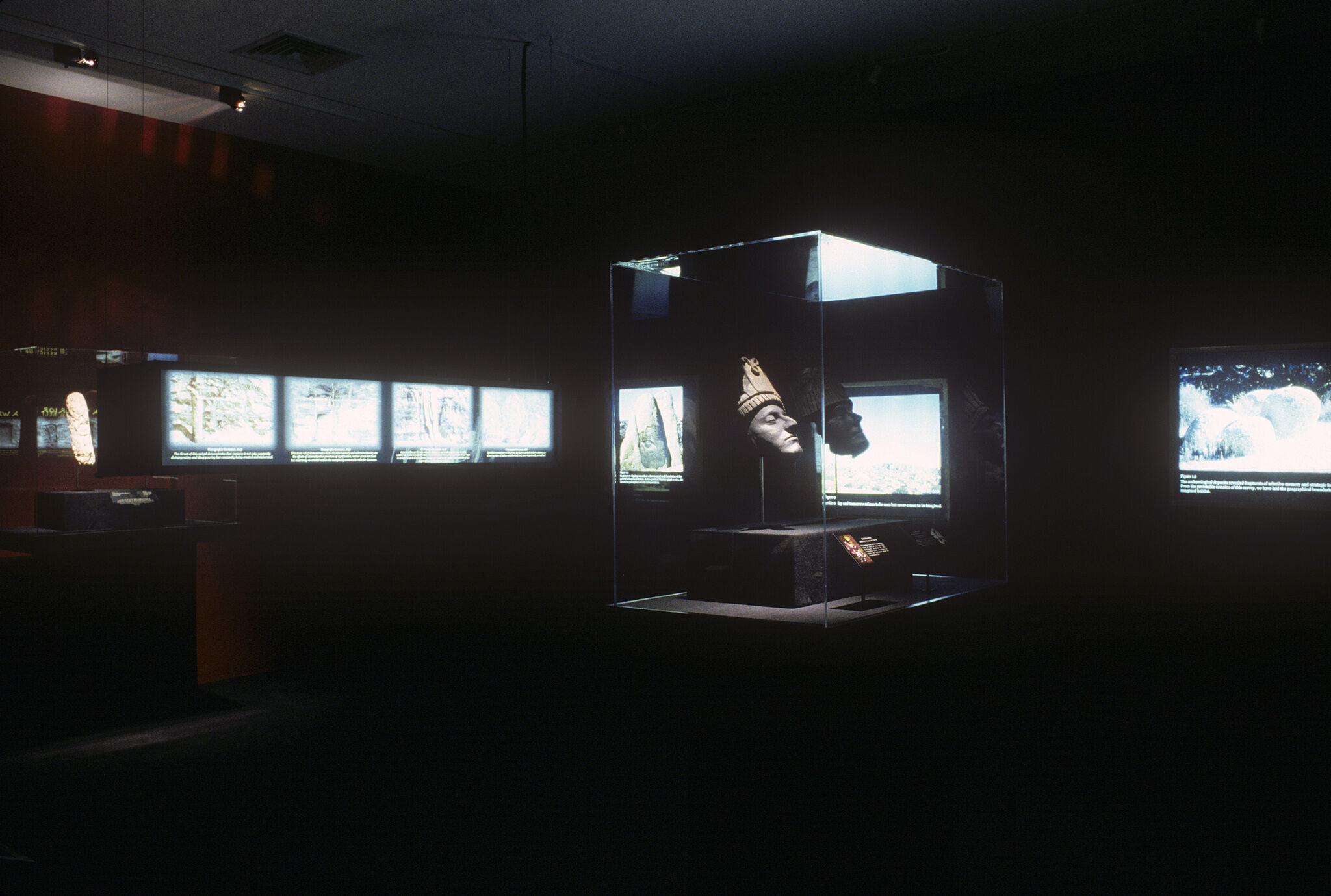 A dark gallery with a sculpture in a glass display case, along with brightly lit monitors around it.