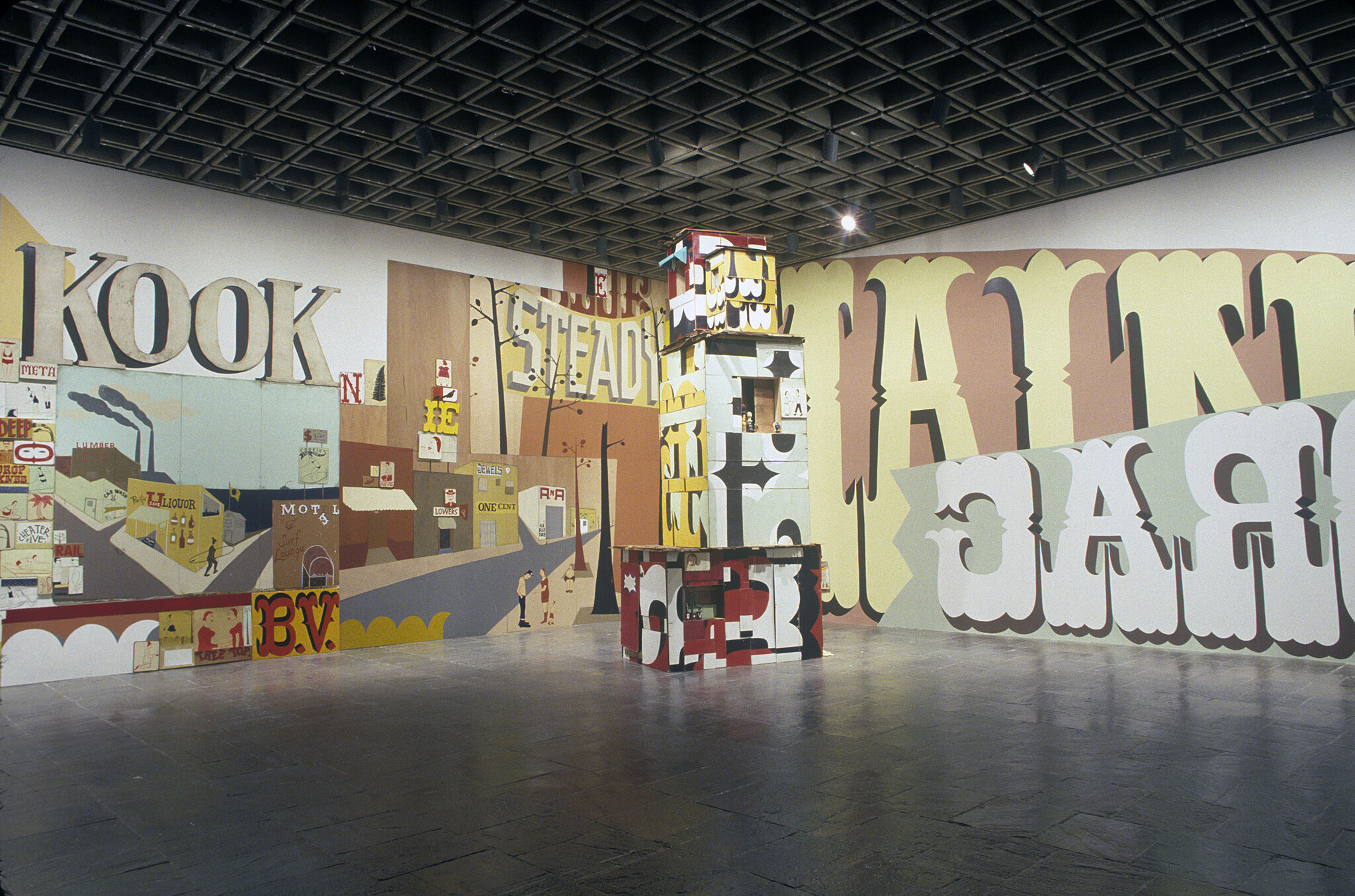 An installation view in a gallery with walls covered in large typography and a sculpture.