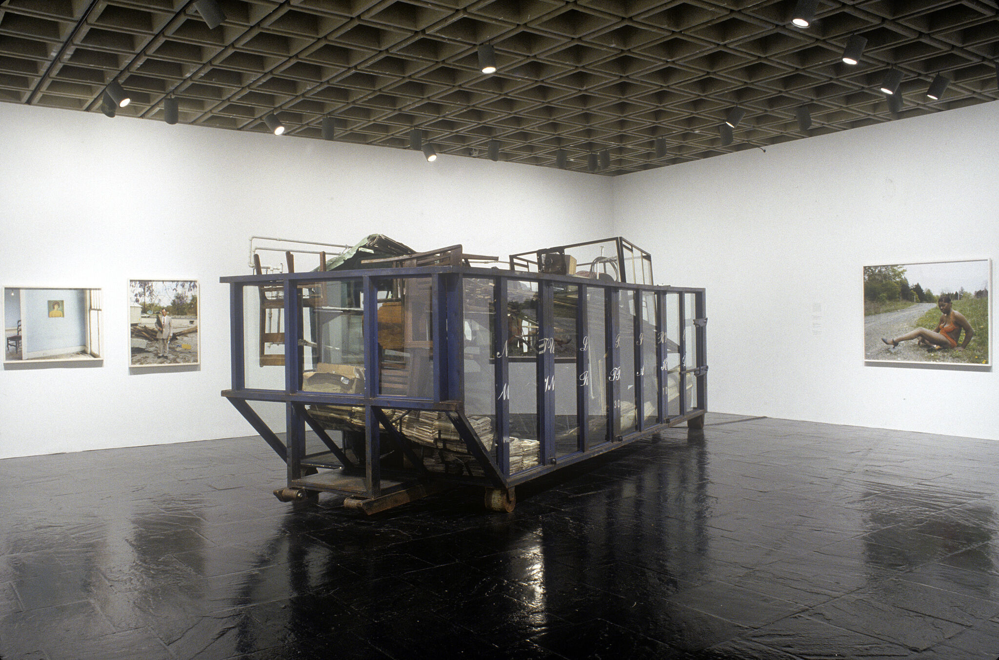 A gallery filled with photographs and a large sculpture of a glass dumpster filled with various objects.