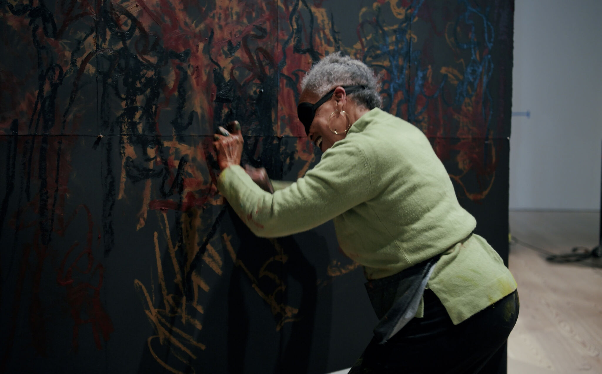 The 2022 Biennial artist Awilda Sterling-Duprey wears a lime green sweater and large gold hoop earrings. She is smiling as she stands blindfolded, pressing against a black wall with intense, jittery, abstract marks on it