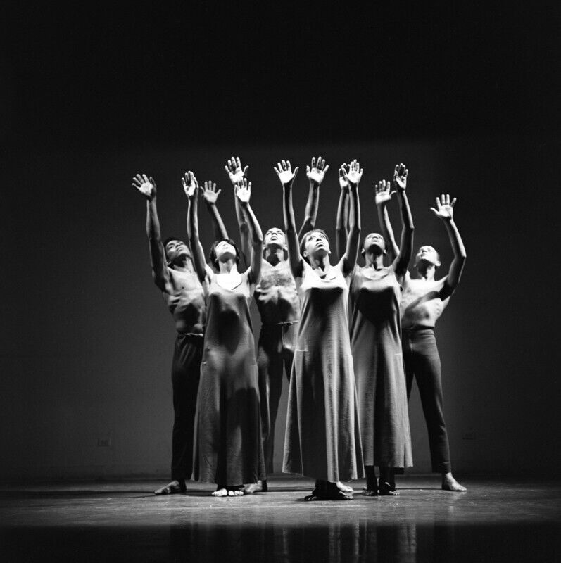 A black- and- white image of a group of dancers standing onstage with their arms stretched towards the ceiling.
