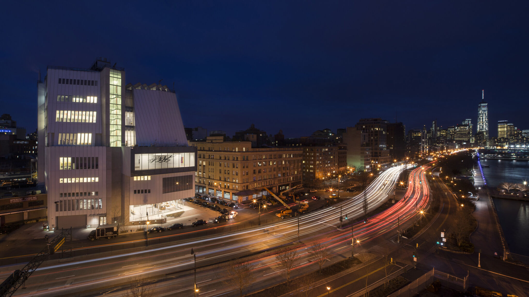Cityscape at night with a brightly lit modern building and streaks of car lights on the road.