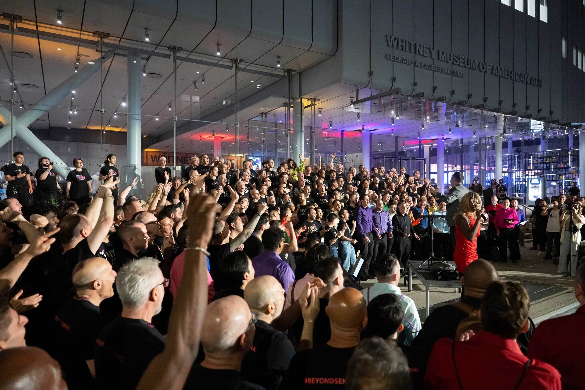Large choir performs outside the Whitney Museum of American Art, with a woman in a red dress speaking.