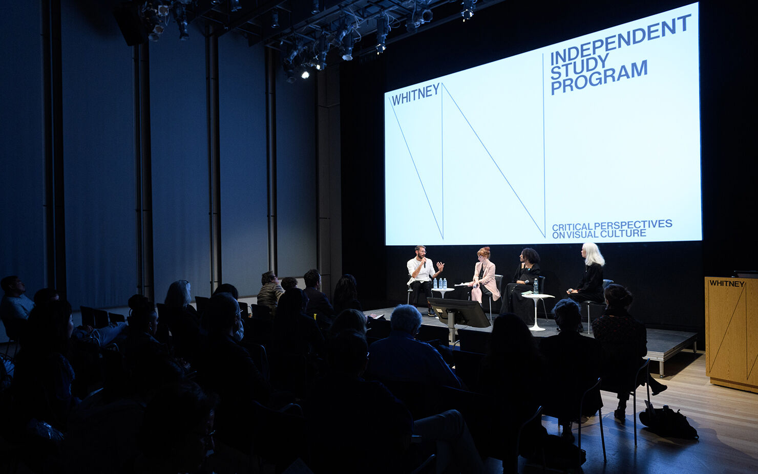 An audience sits in front of a panel discussion. There is a projected slide that reads Whitney Independent Study Program.