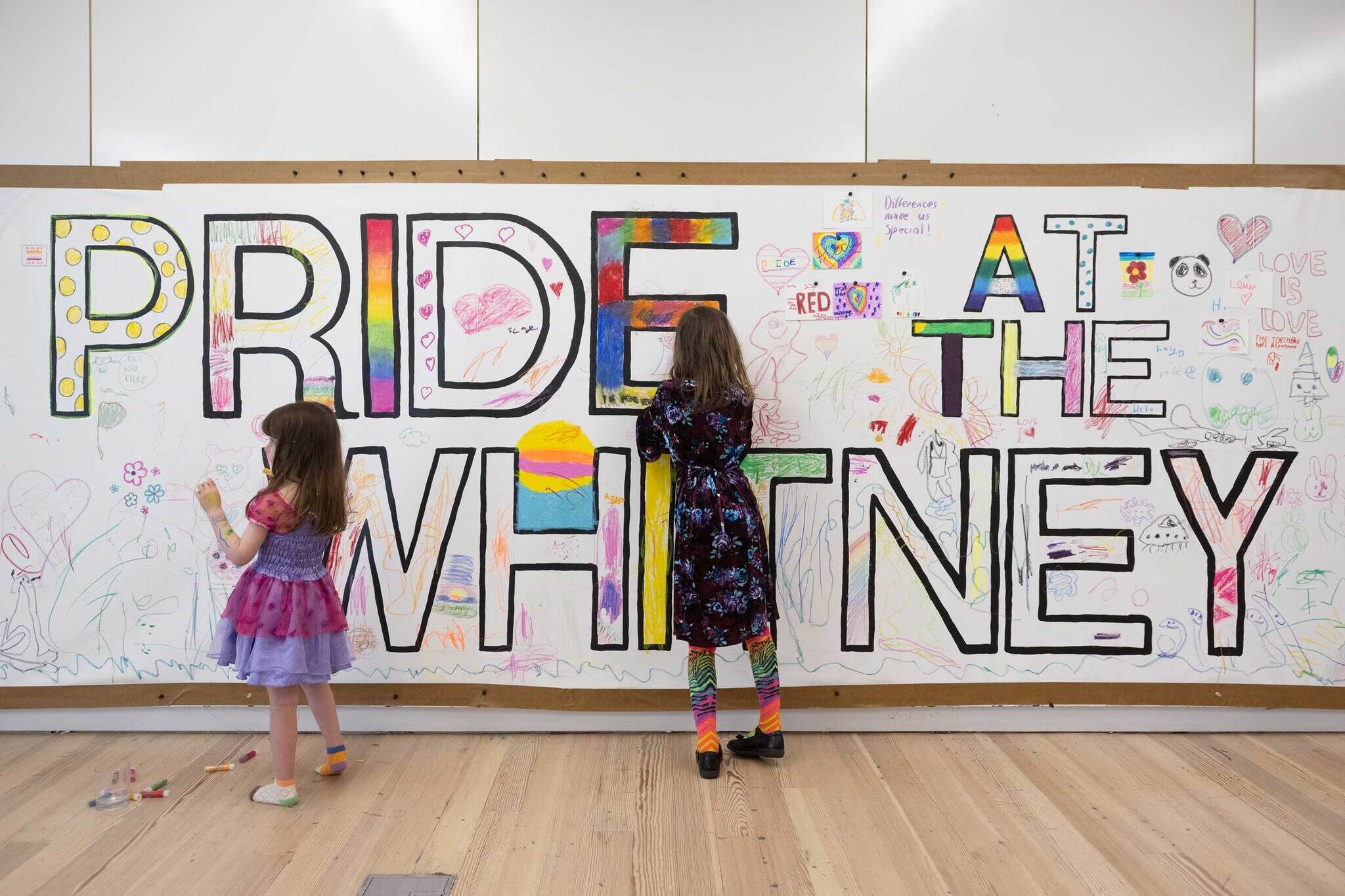 Two children color and draw on a large mural that says “PRIDE AT THE WHITNEY” with rainbow designs.
