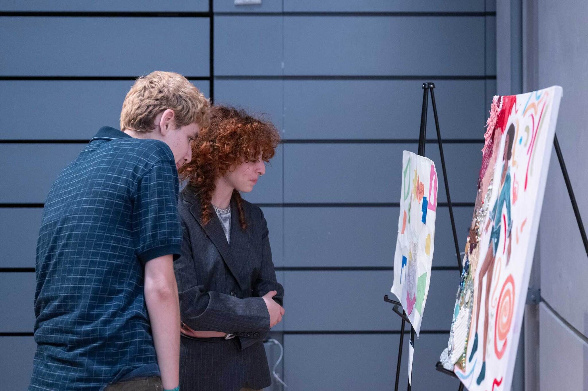 Two people closely examine colorful artwork displayed on easels in a modern indoor setting.