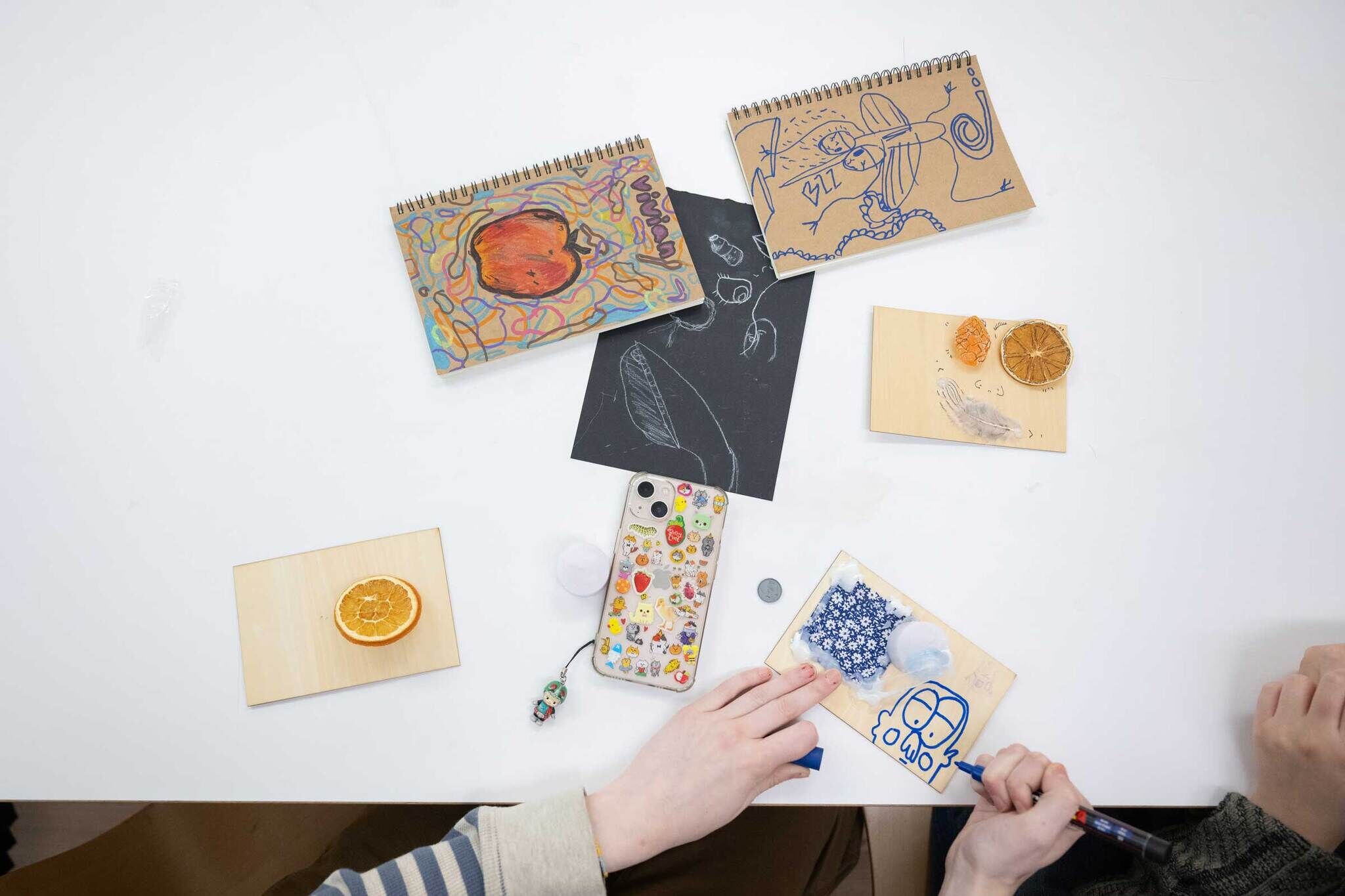 On a table sits multiple wooden flat rectangular panels with an assemblage of materials on top for a collage.