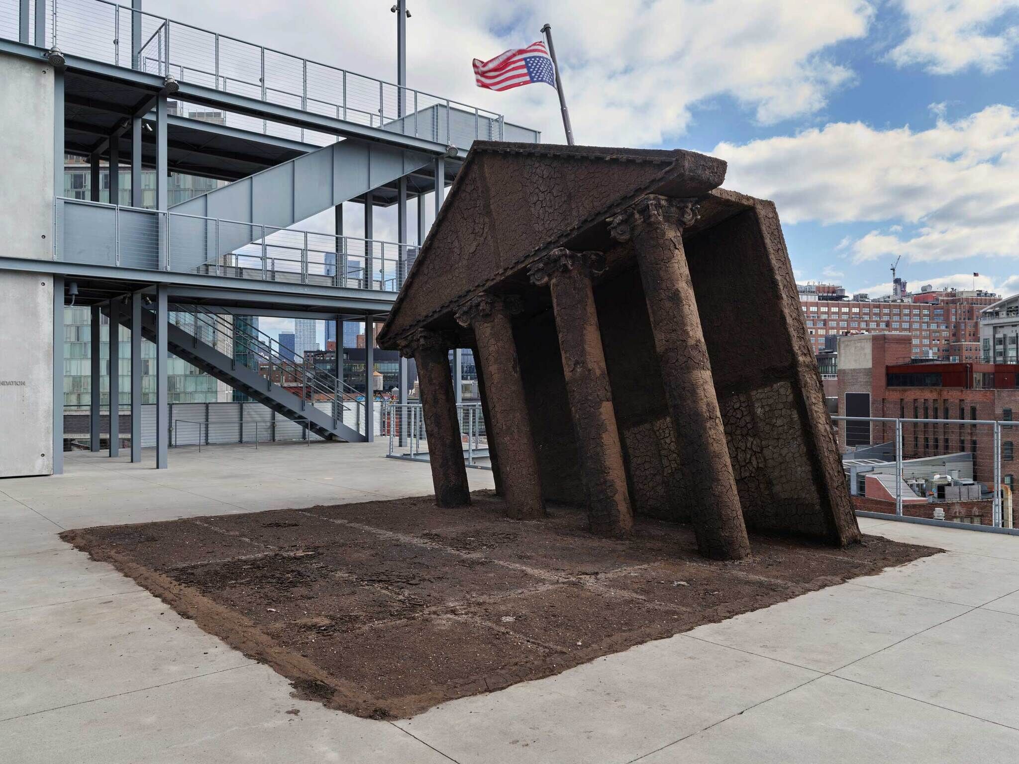A weathered, upside-down building facade that looks like the White House installed on an urban plaza with a staircase and American flag in the background.