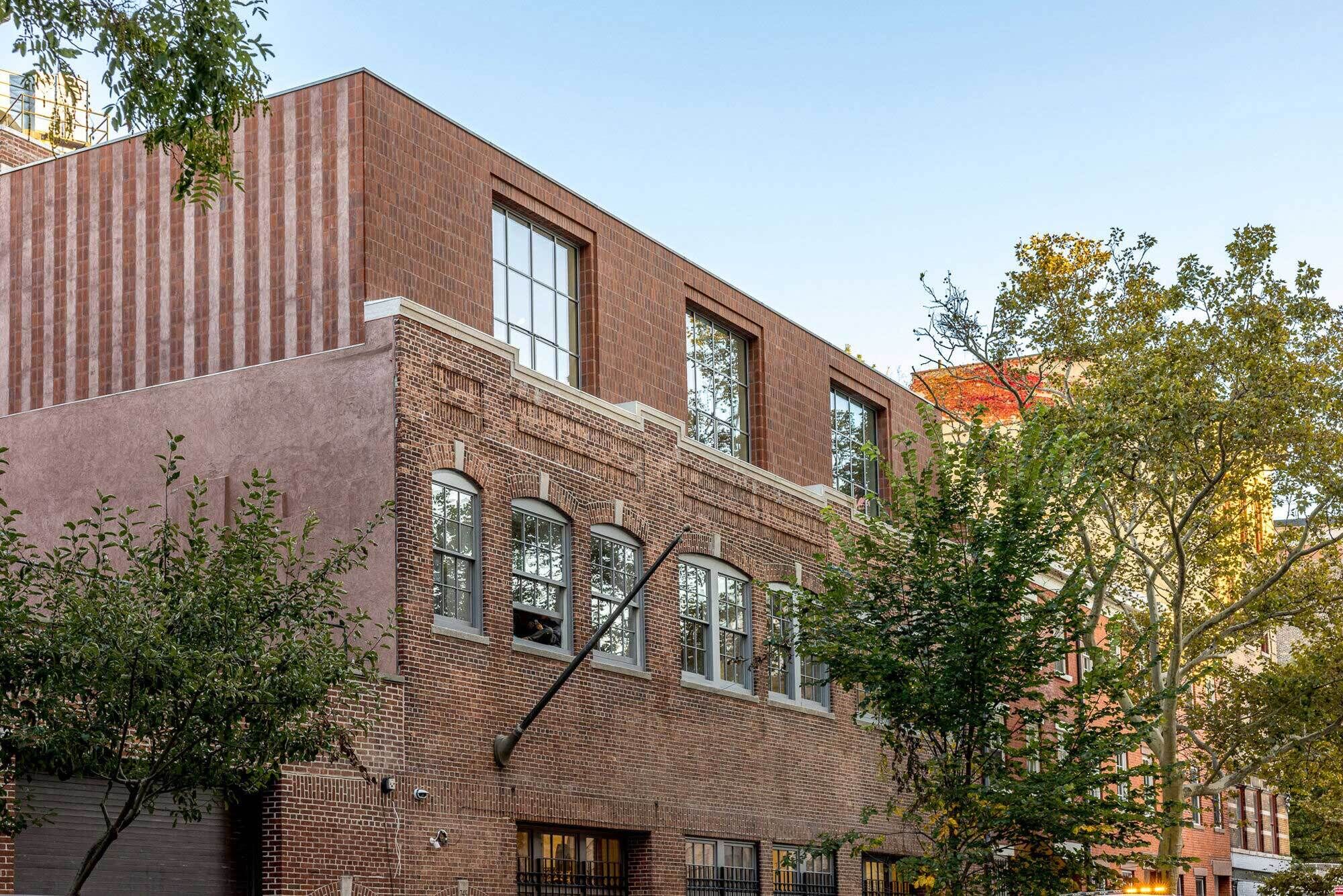 A three-storied brick building with large front facing windows with trees in the foreground and blue sky.  