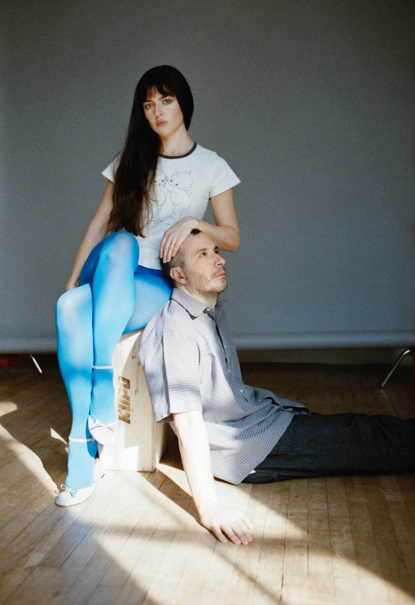 A woman in blue tights sits on a box, resting her hand on a man’s head as he sits below.