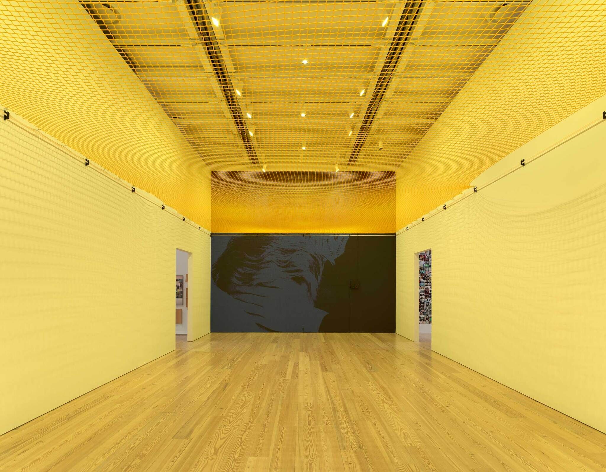 Empty gallery room awash with yellow walls, wooden floor, and a large abstract painting on the back wall.