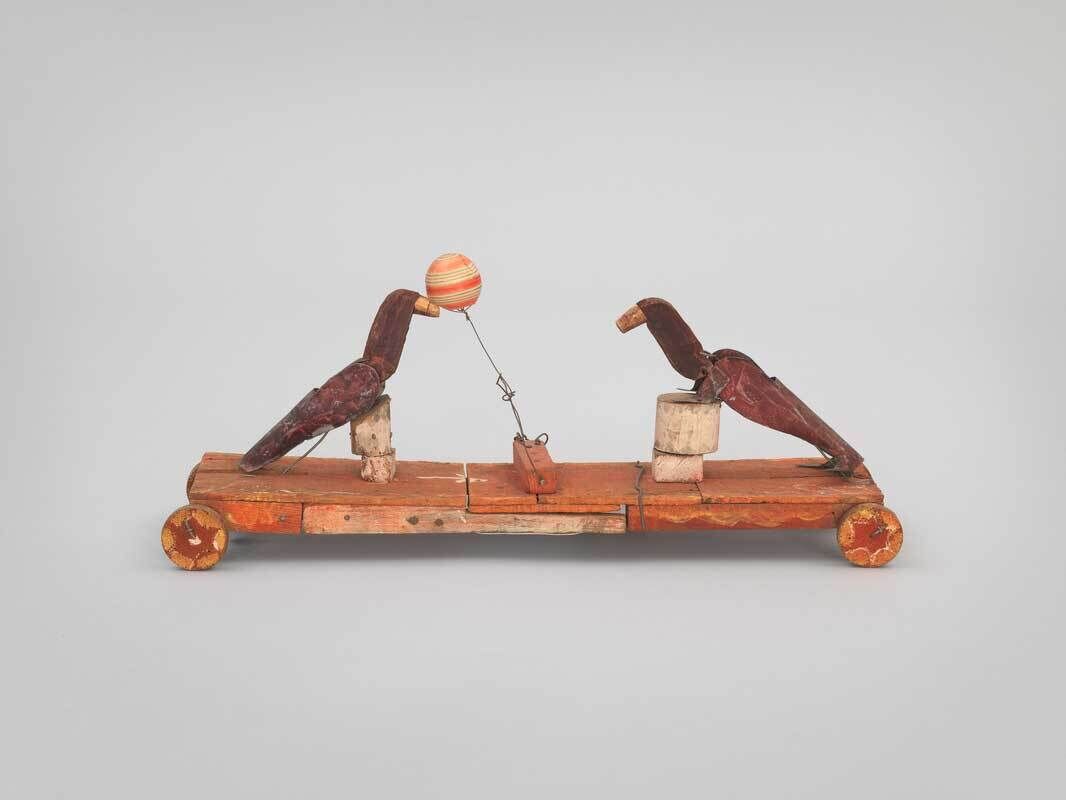 Image of a wooden figurine of two dark brown seals balanced on white pedestals atop a red-brown rolling platform. The seals are poised to pass an orange and white ball mounted between them on a piece of wire.