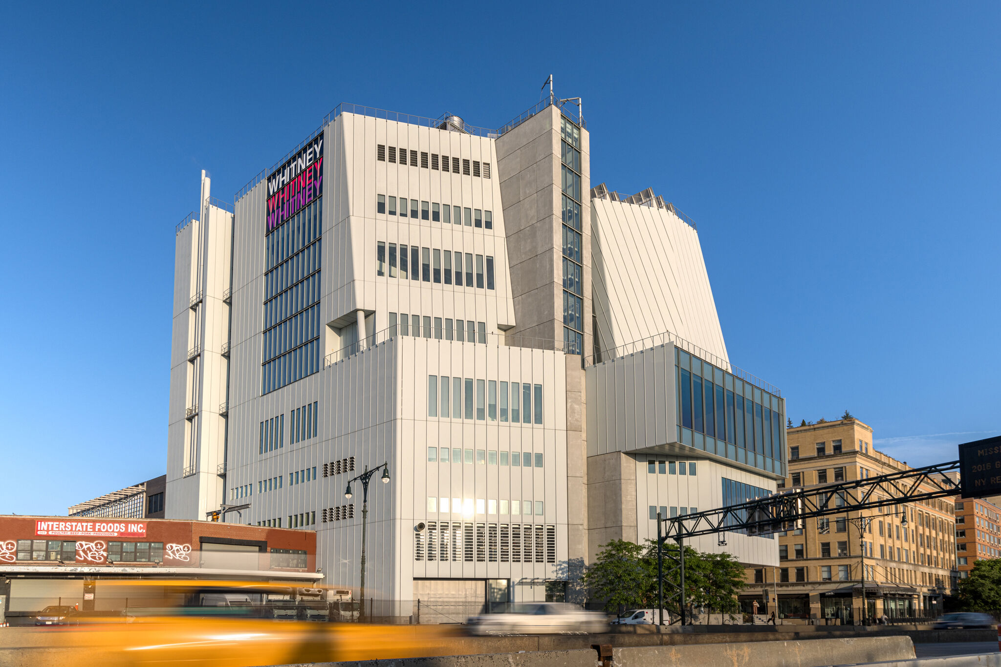 Large modern building with “Whitney” sign in bold letters, yellow taxi passing by on the street below.