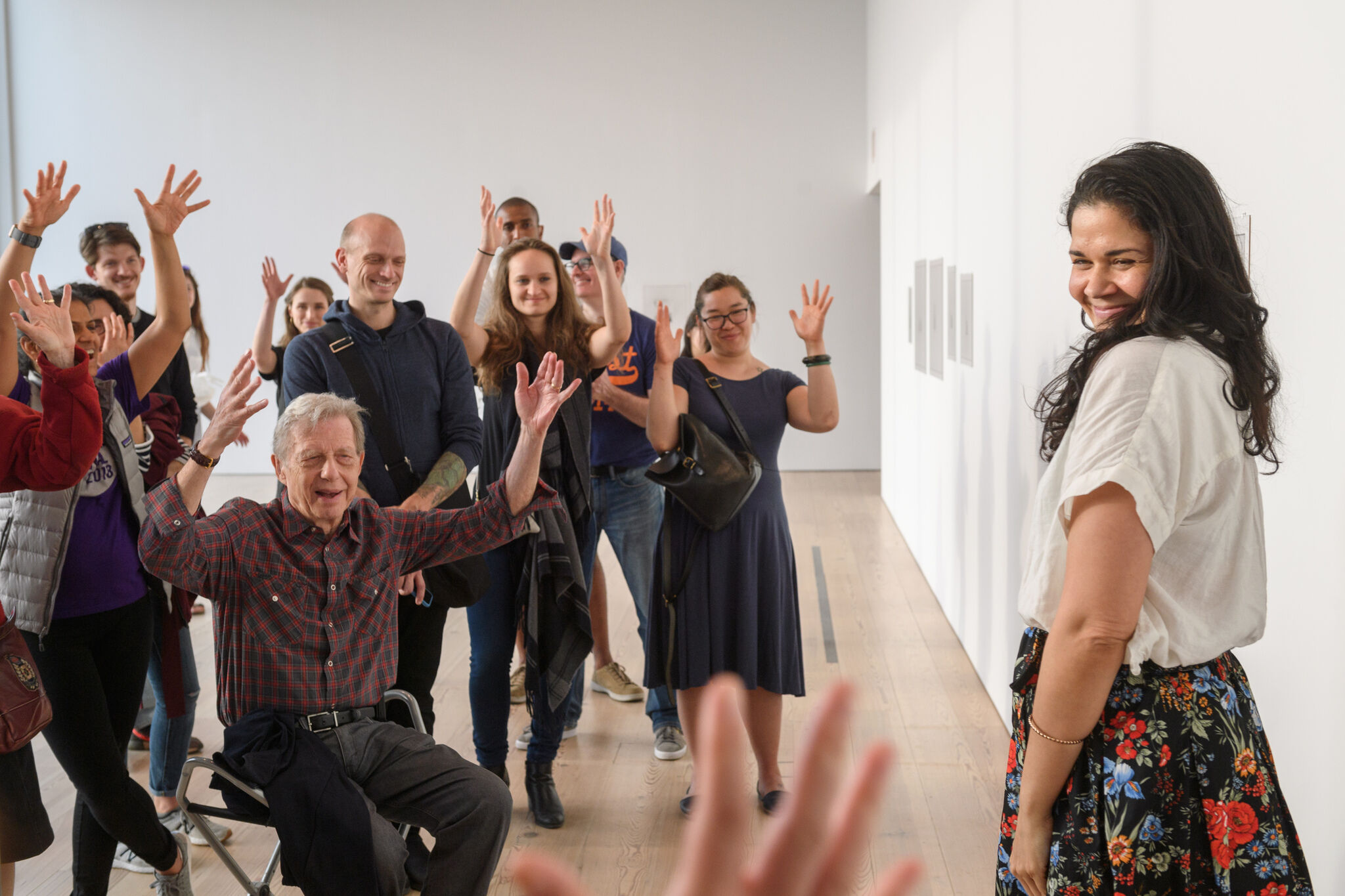 A group of people, some smiling and raising their hands, face a woman who is also smiling.