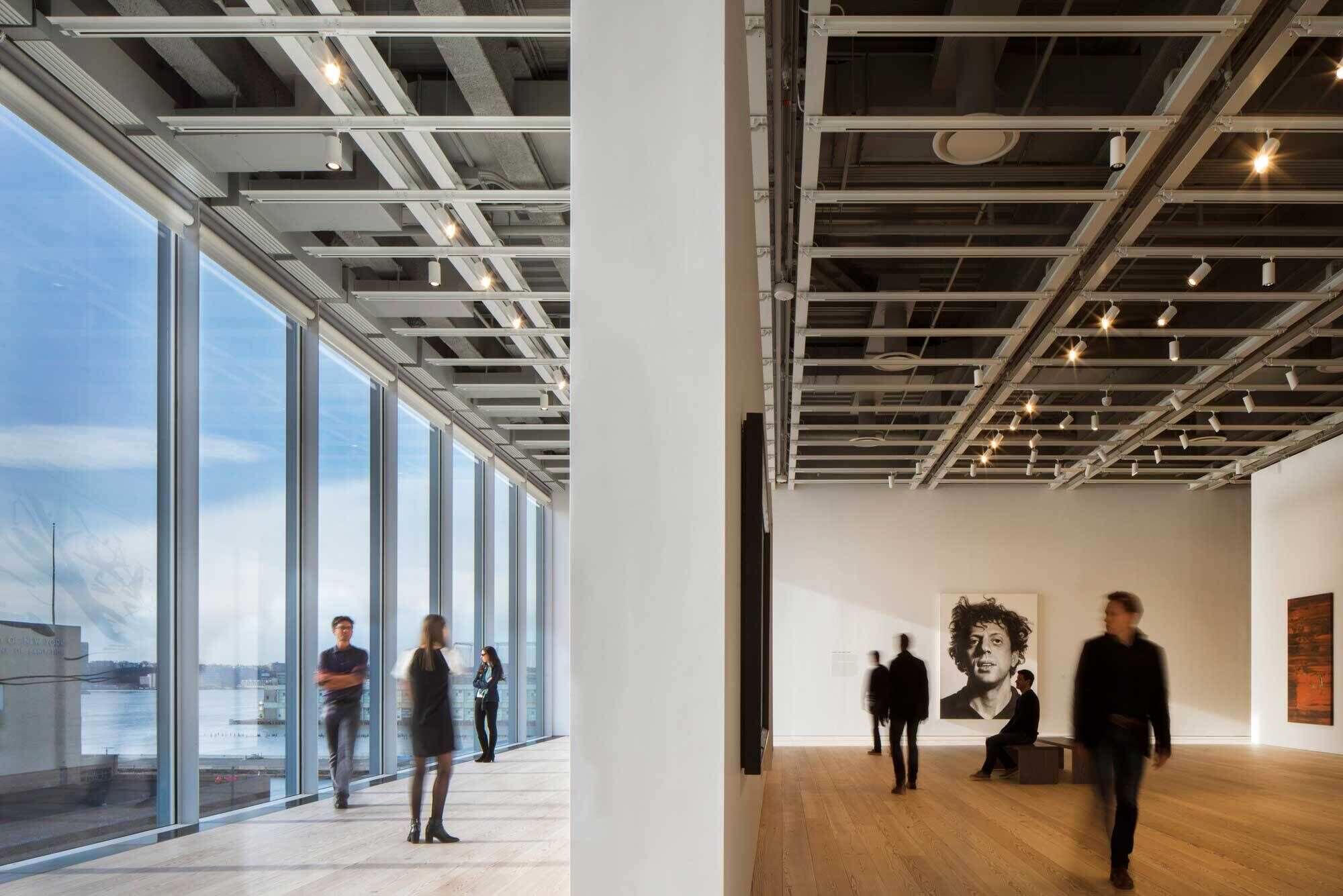 People walk and stand inside a modern art gallery with large windows and a black-and-white portrait on the wall.