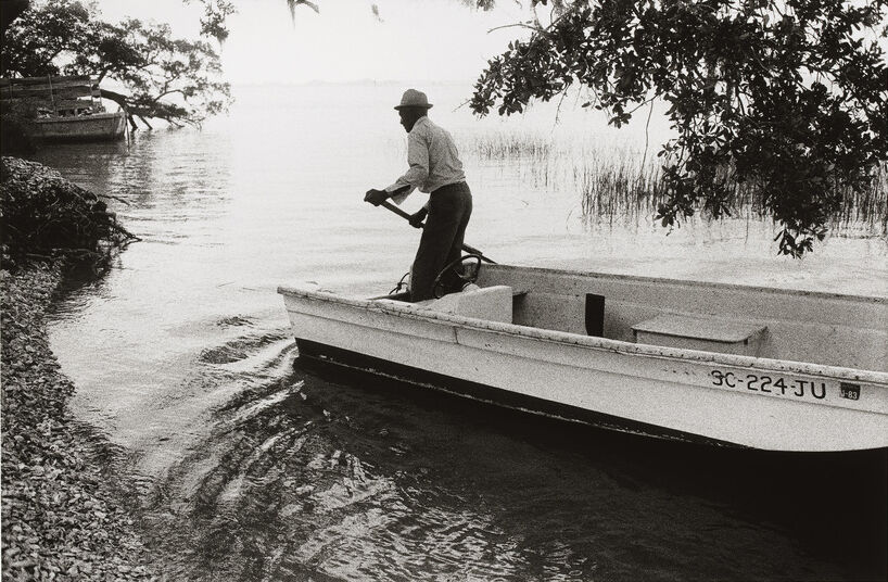 A person in a hat steers a small boat near a shoreline with trees and reeds, creating a serene, black-and-white scene.