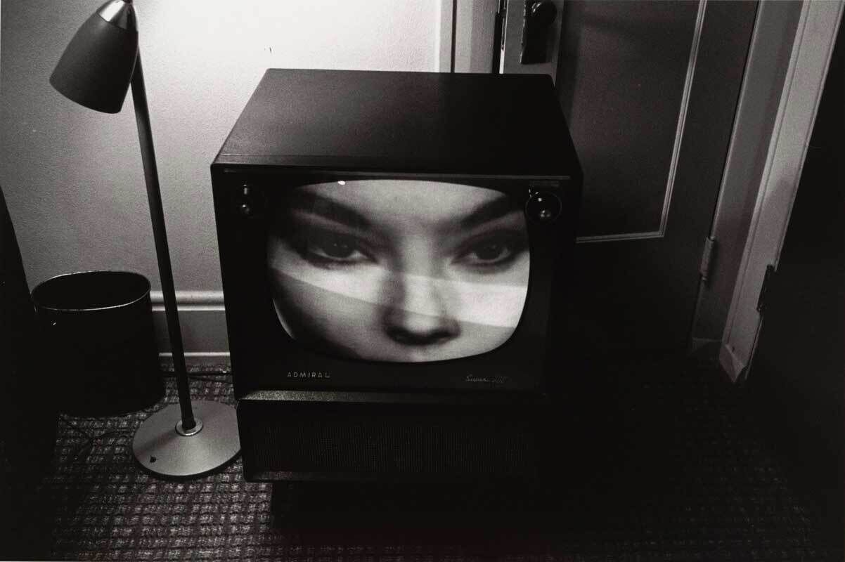 A black and white image of a tv on the floor in a hotel room next to a lamp with a close up on a woman's face. 