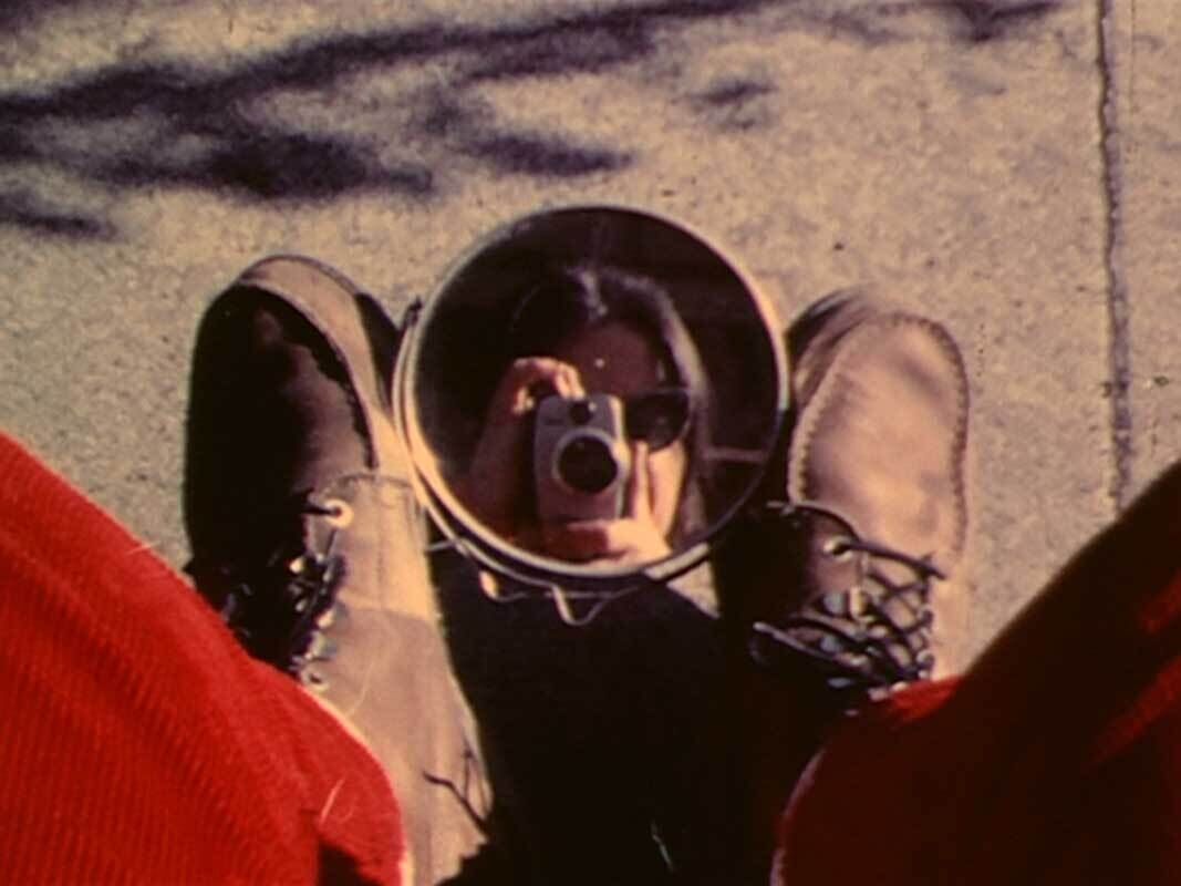 A woman takes a portrait of herself with a camera, framed in a mirror positioned on the ground between her feet.