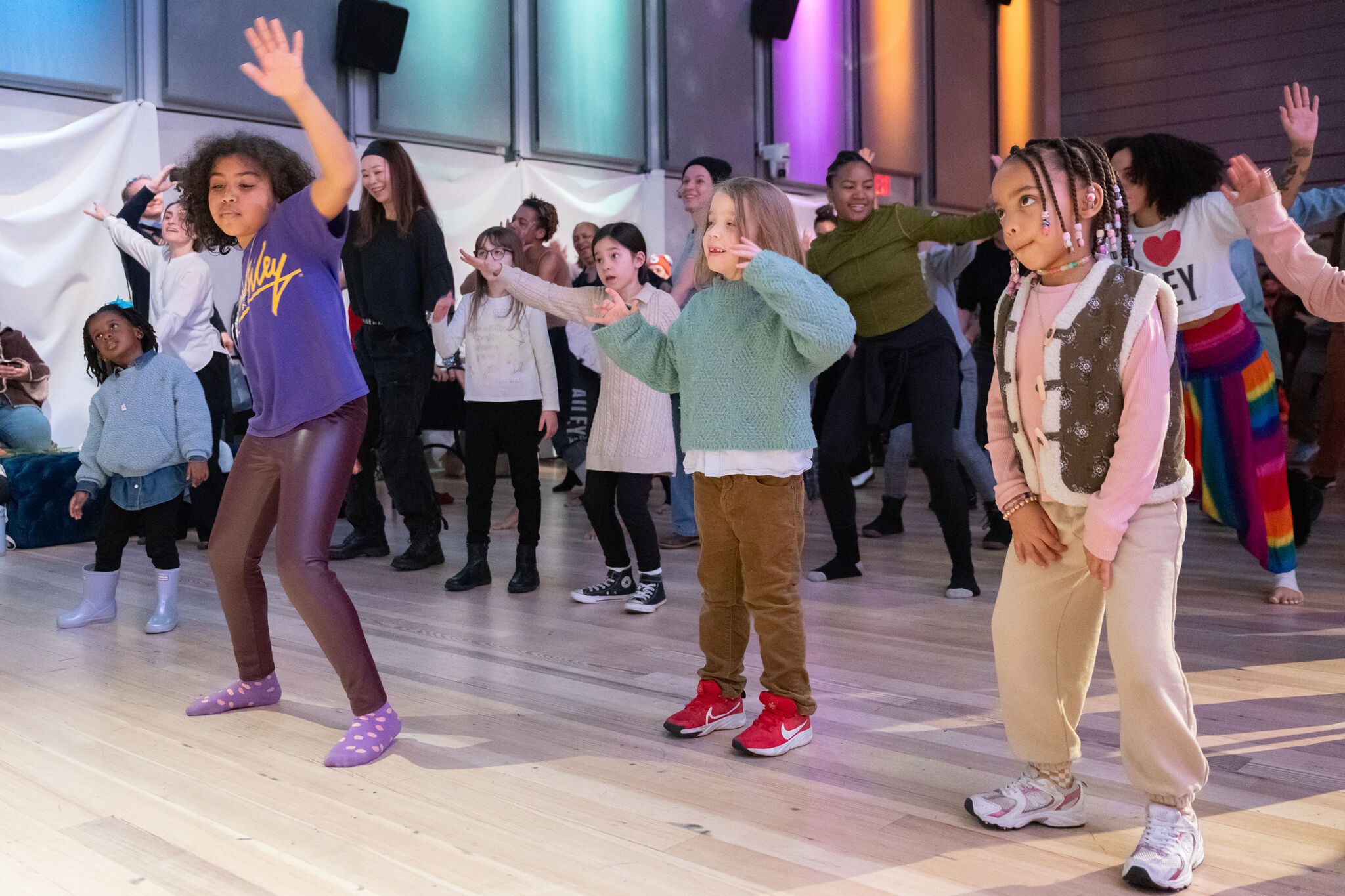 Children and adults dance together in a brightly lit room, following the same energetic dance moves.