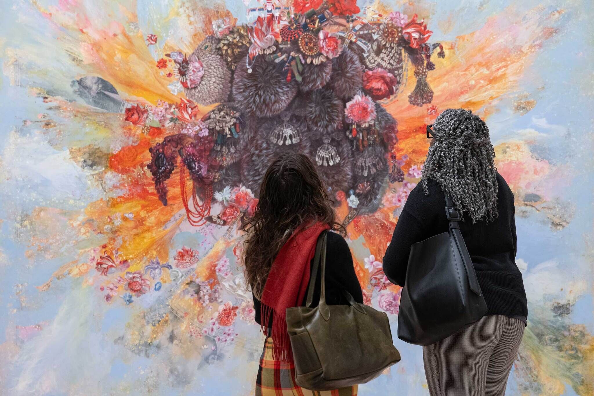 Two people with bags stand closely together, looking at a large, colorful abstract painting with flowers and textures.