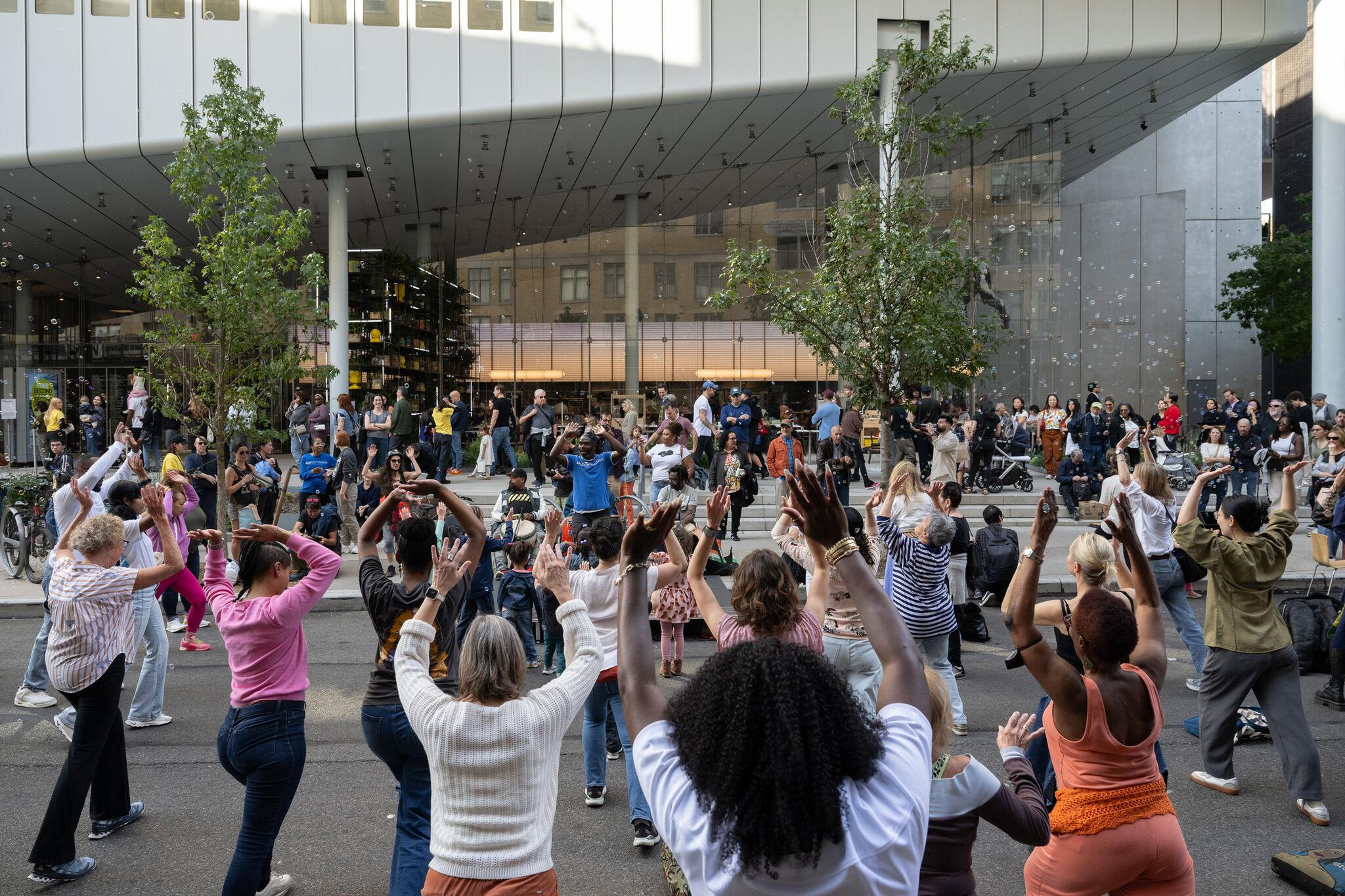 A large group of people dance together outdoors, raising their arms, while others watch and walk nearby.