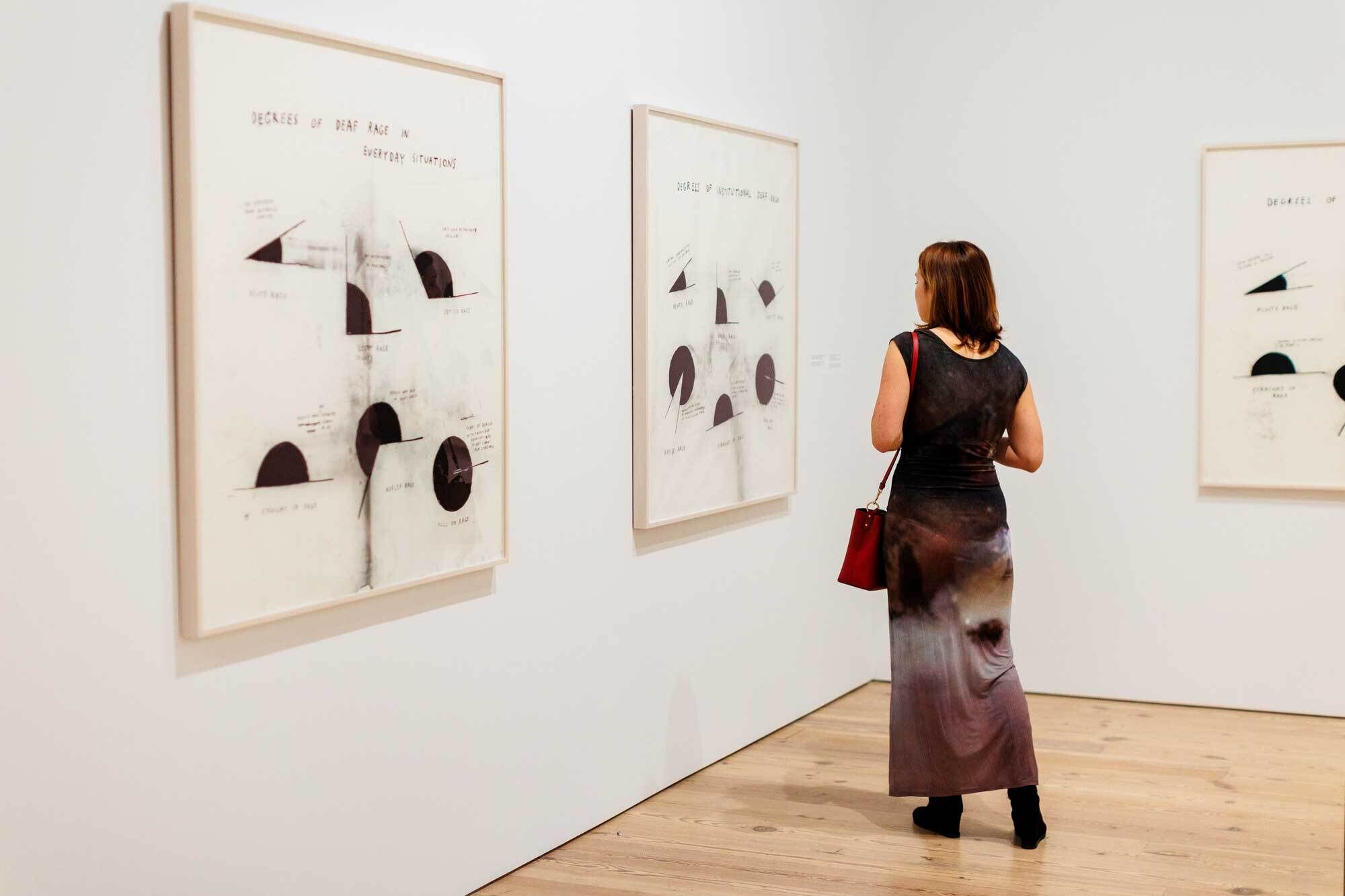 A woman with a red purse looks at framed abstract art on a white gallery wall.