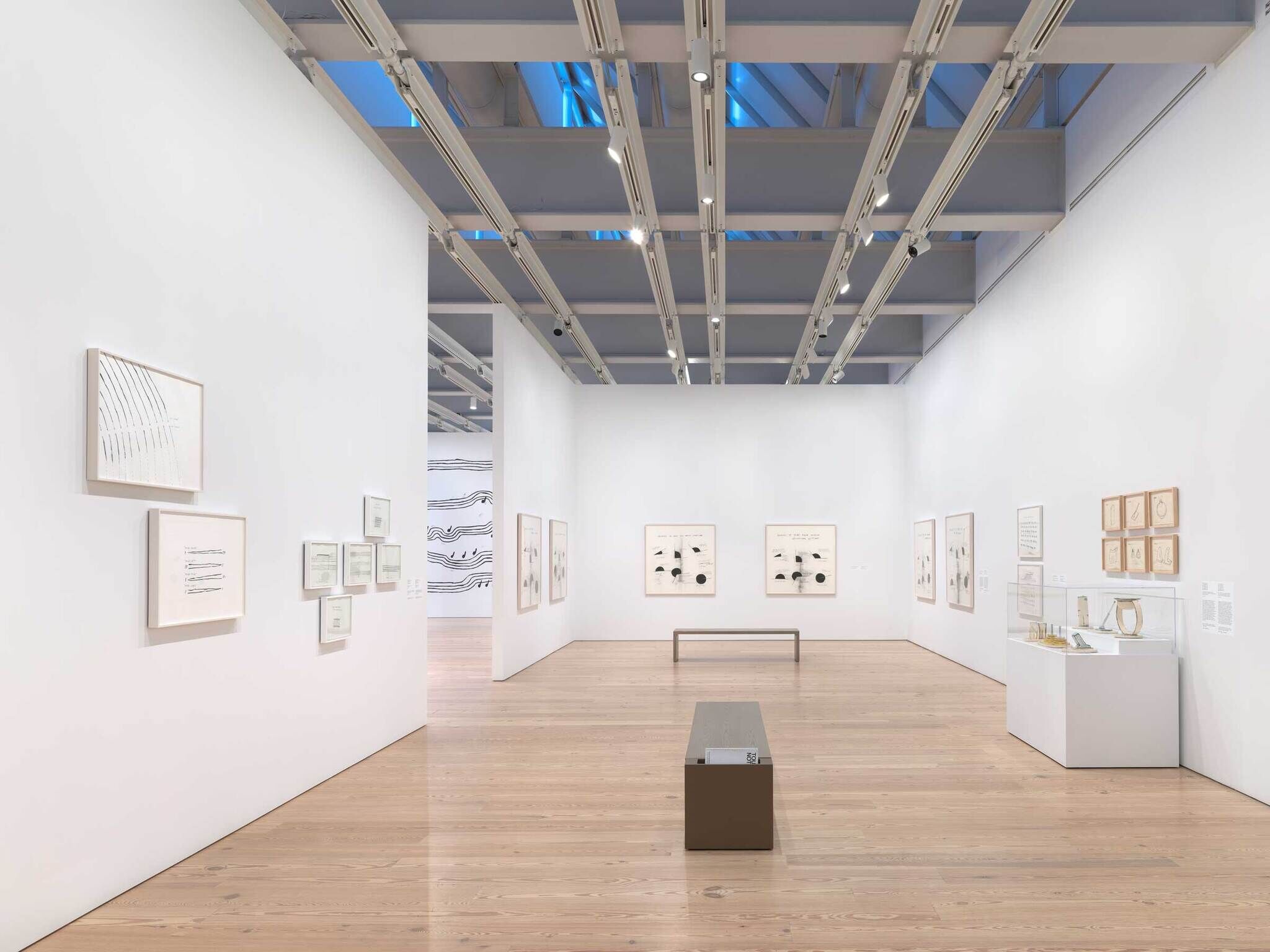 Spacious art gallery with framed drawings on white walls, wooden floor, and a central bench.