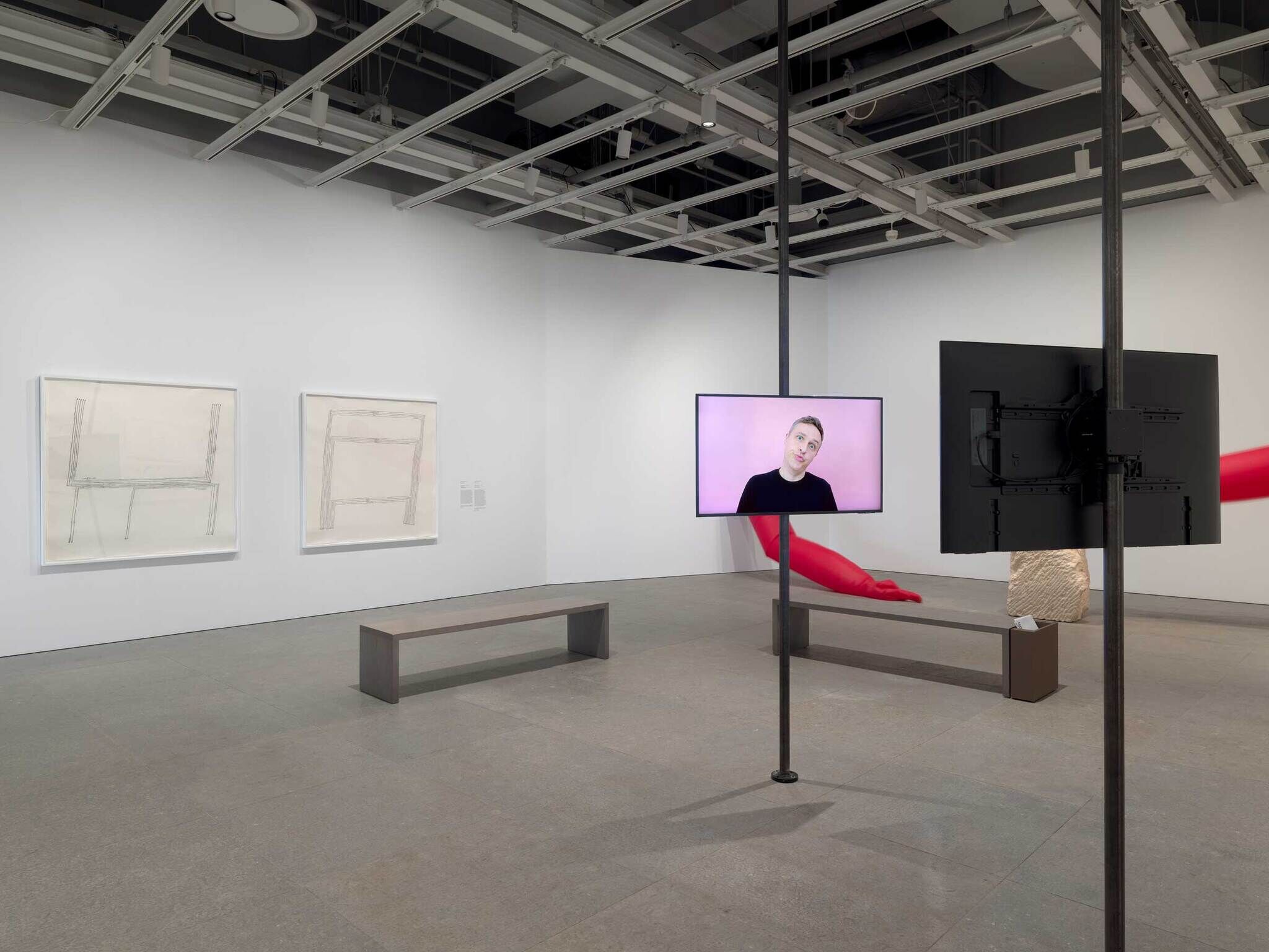 Art gallery with two framed sketches, a video screen showing a person, and a red sculpture.