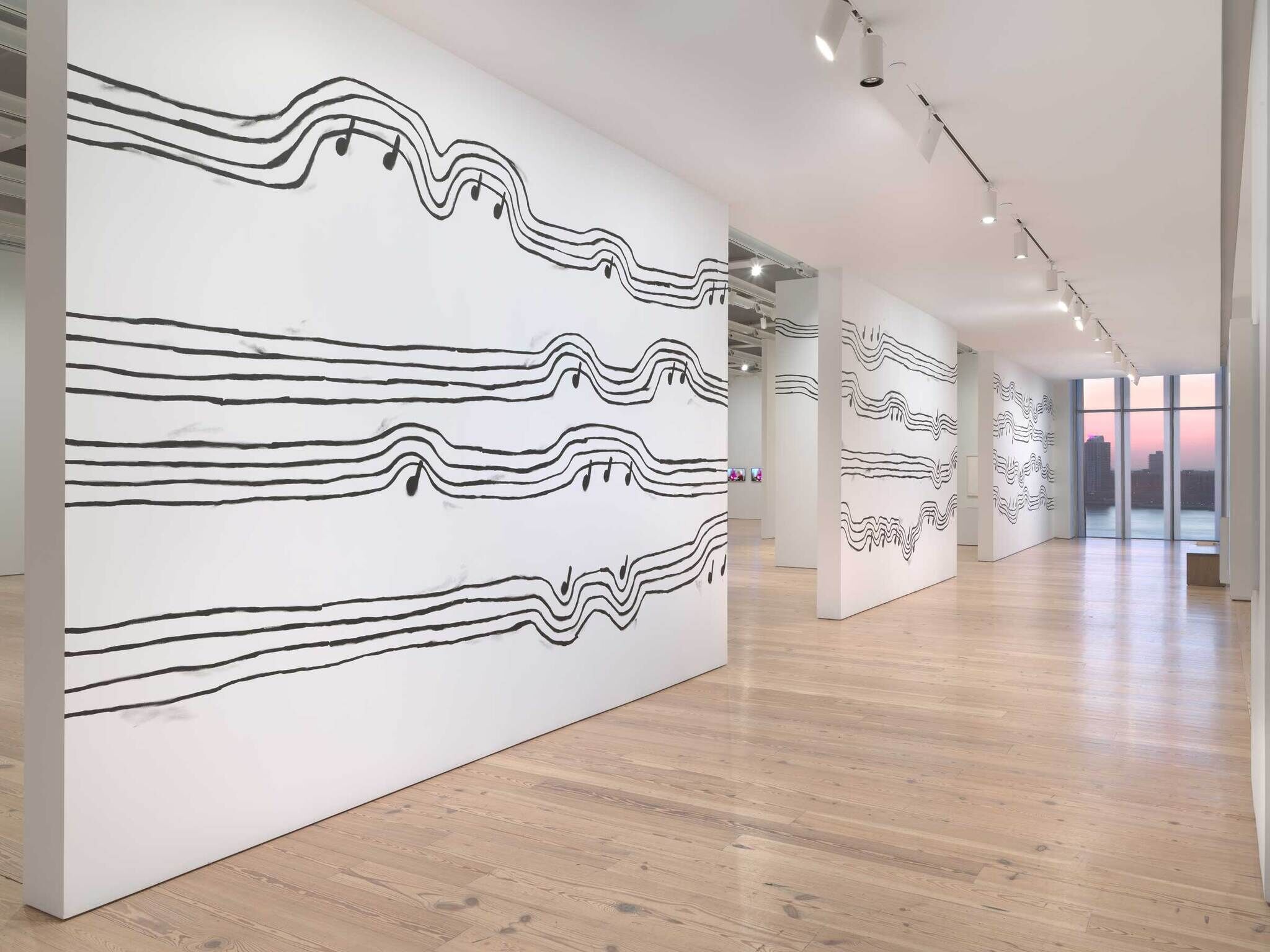 Art gallery with large white walls featuring black wavy lines and musical notes, wooden floor, sunset view.