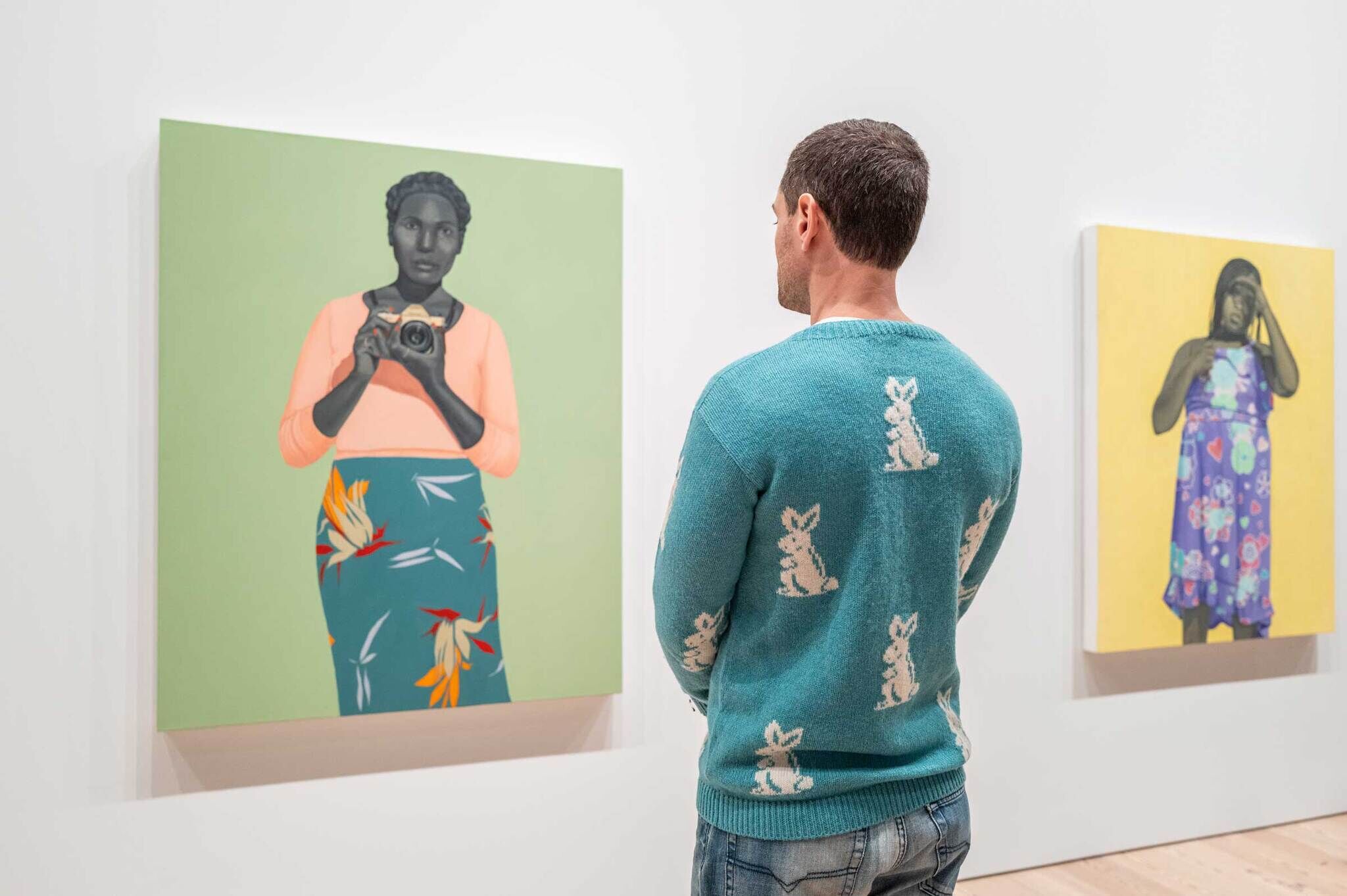 A young man in a blue sweater with white print on it is looking at a painting of a young woman with a camera in her hand gazing back at the viewer.