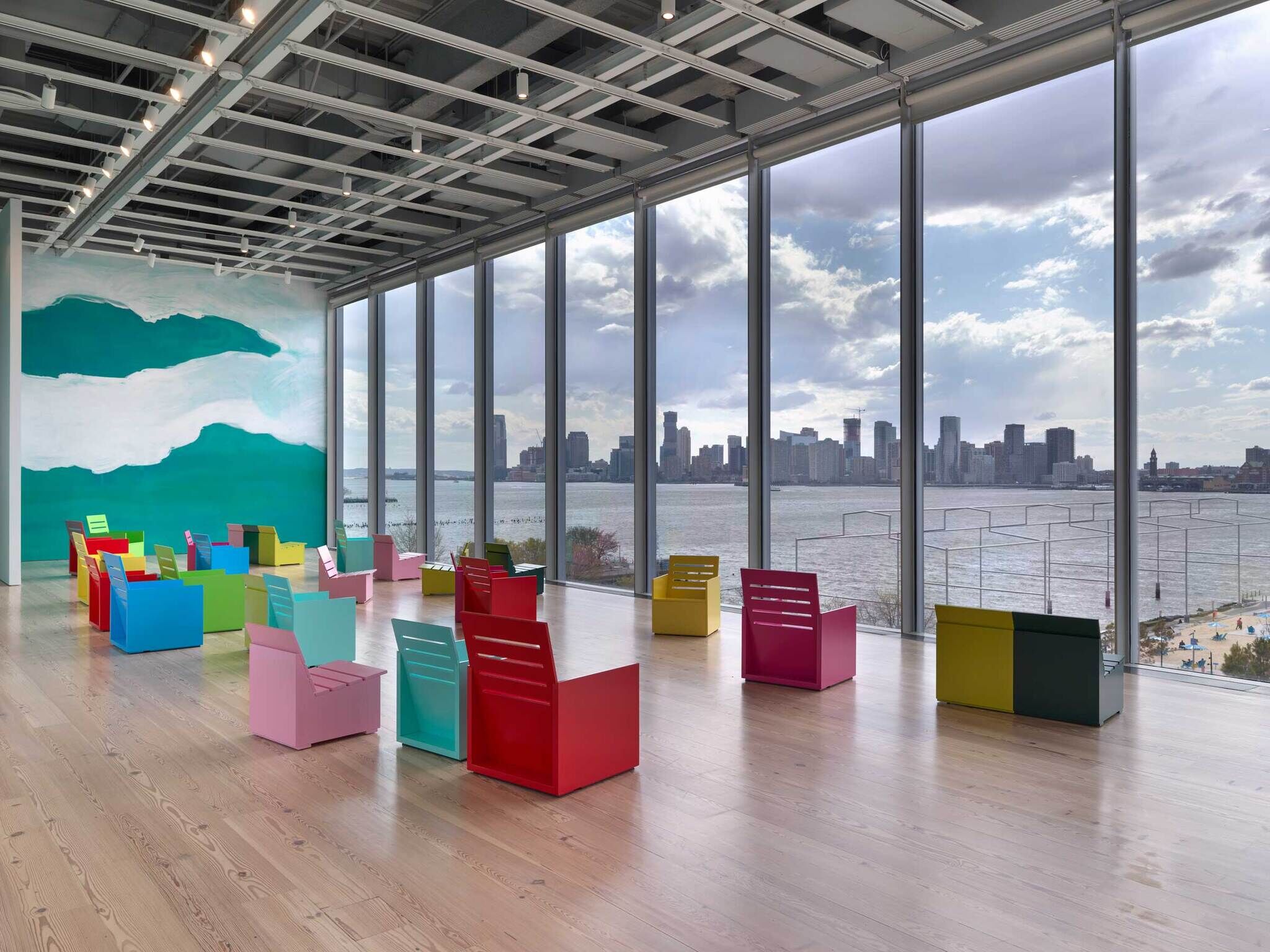 Brightly colored chairs are arranged in a spacious room with large windows overlooking a city skyline and river.