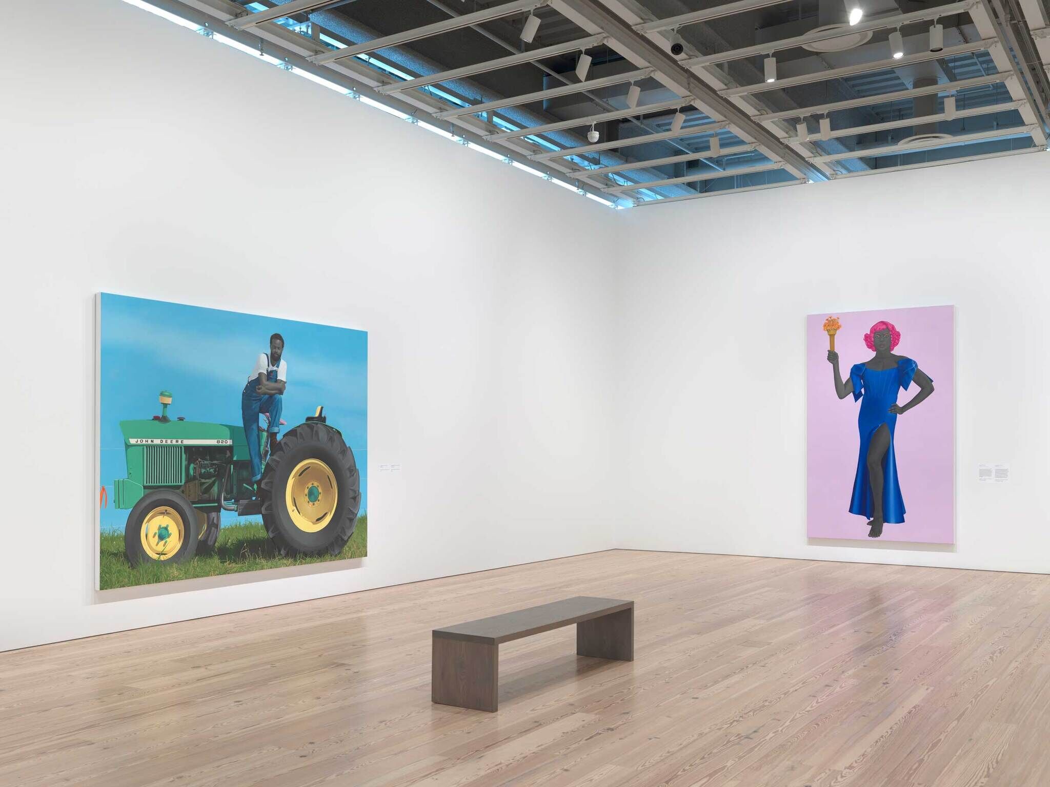 Two large colorful paintings hang in a gallery; one shows a person on a green tractor, the other a person in a blue dress holding a torch.