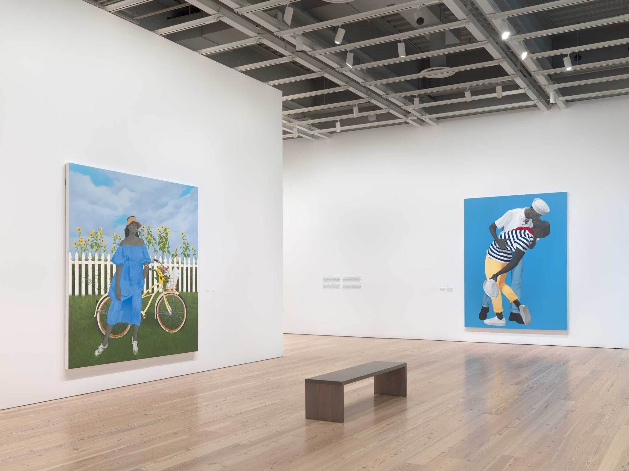 Two large colorful paintings hang on white gallery walls, with a wooden bench placed on the floor between them.