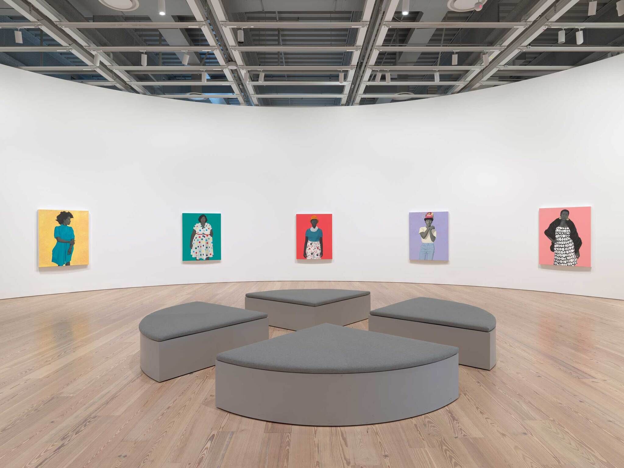 Six colorful portraits of people hang on a curved white wall, with gray benches arranged in the center.