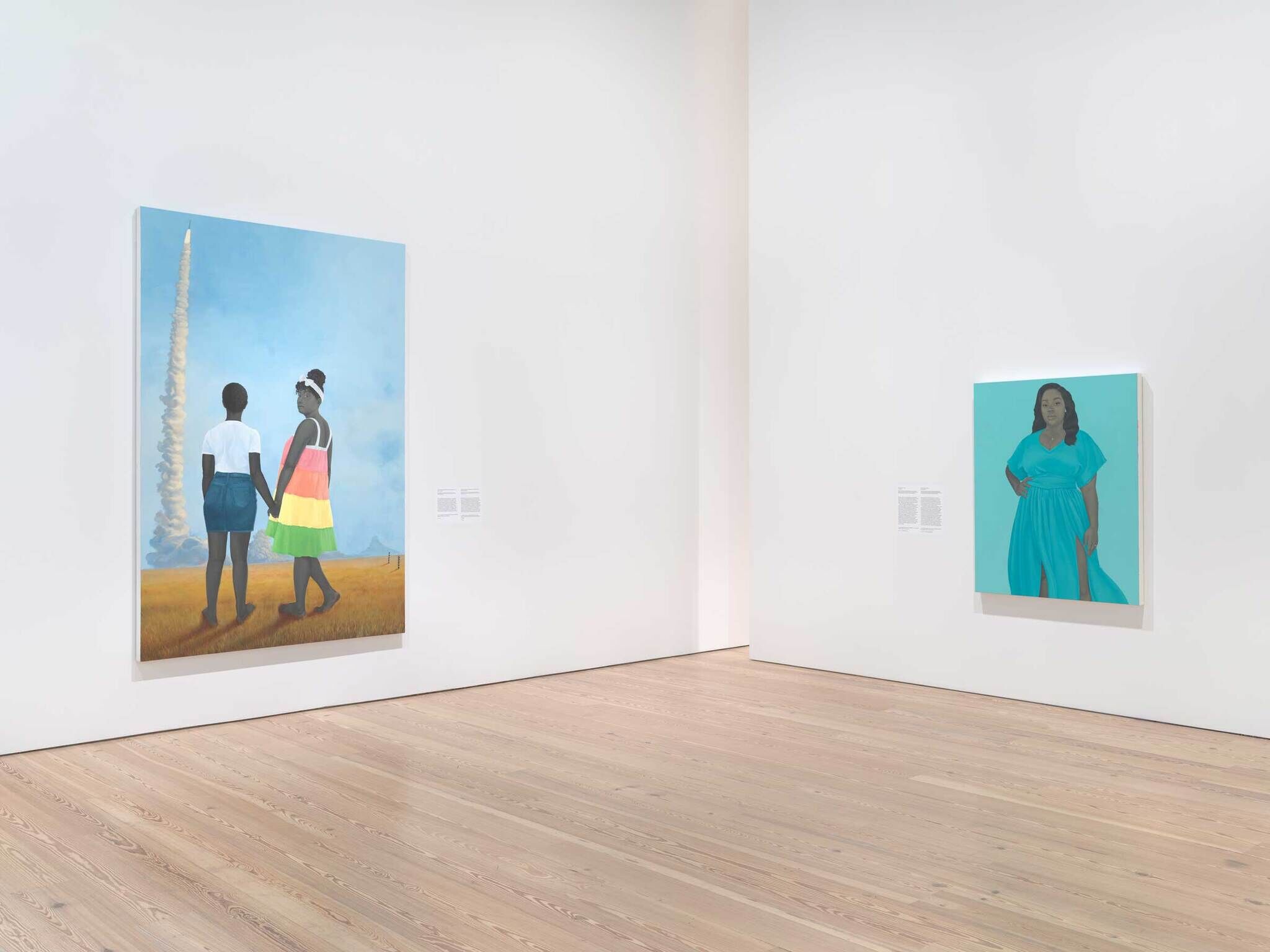 Two colorful paintings hang on a white gallery wall; one shows two people holding hands, the other a woman in blue.