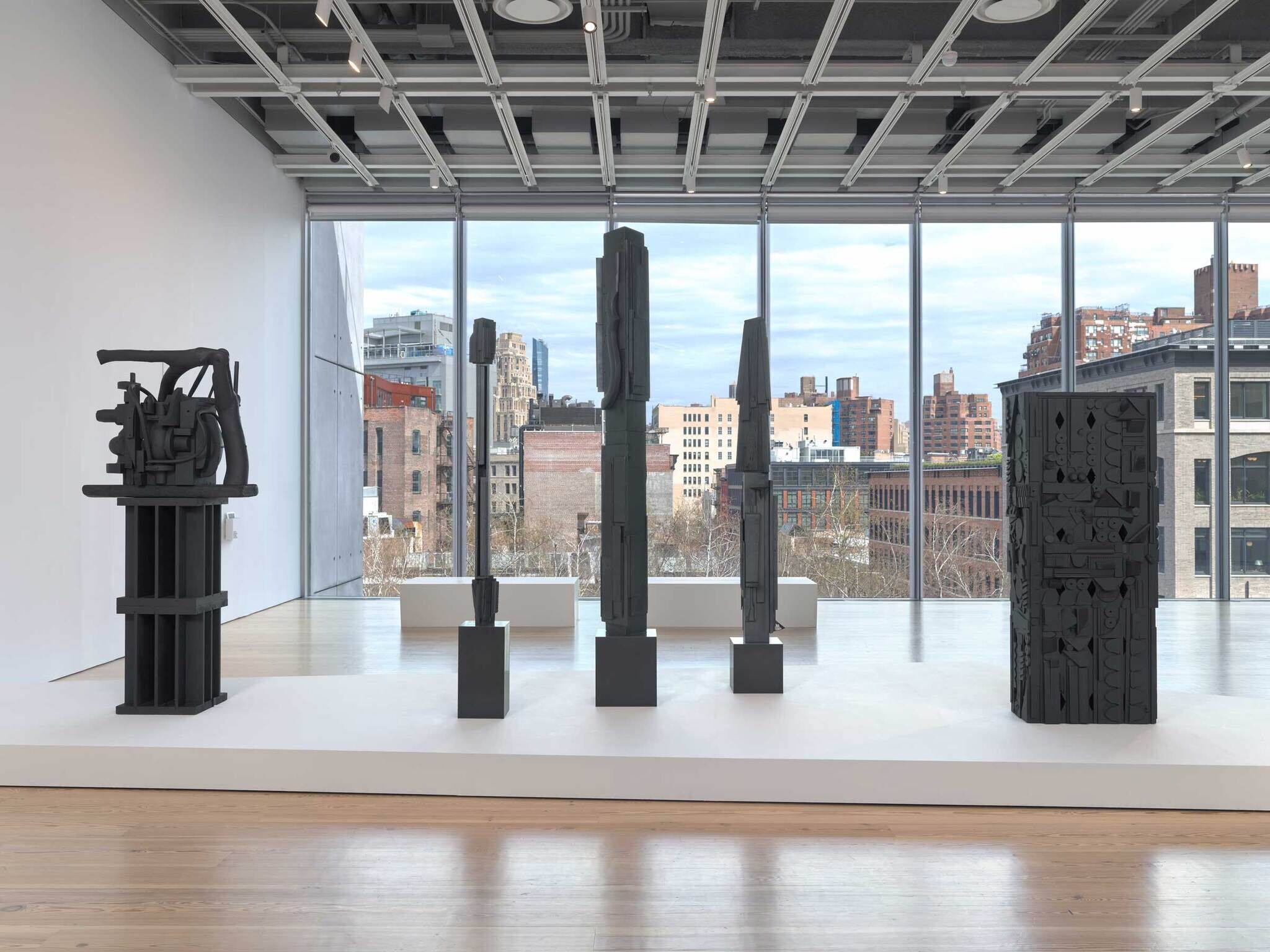 Five tall, dark abstract sculptures stand on a white platform in a bright gallery with large windows.