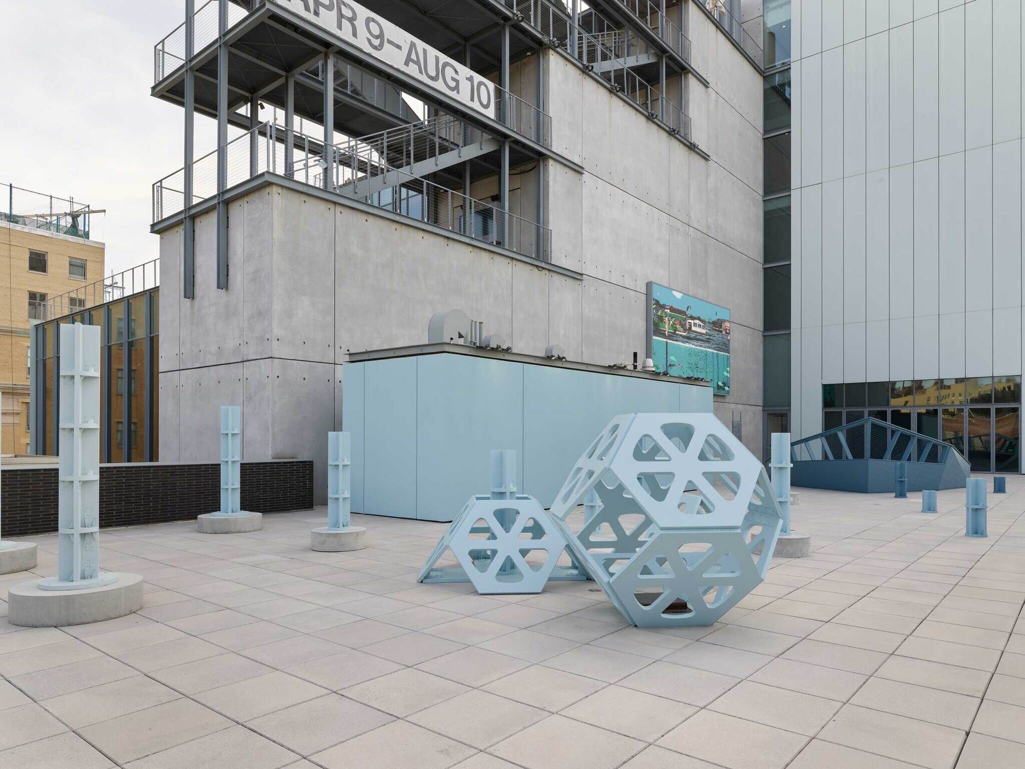 Outdoor terrace with large light blue geometric sculptures, concrete pillars, and a sign reading "APR 9 - AUG 10."
