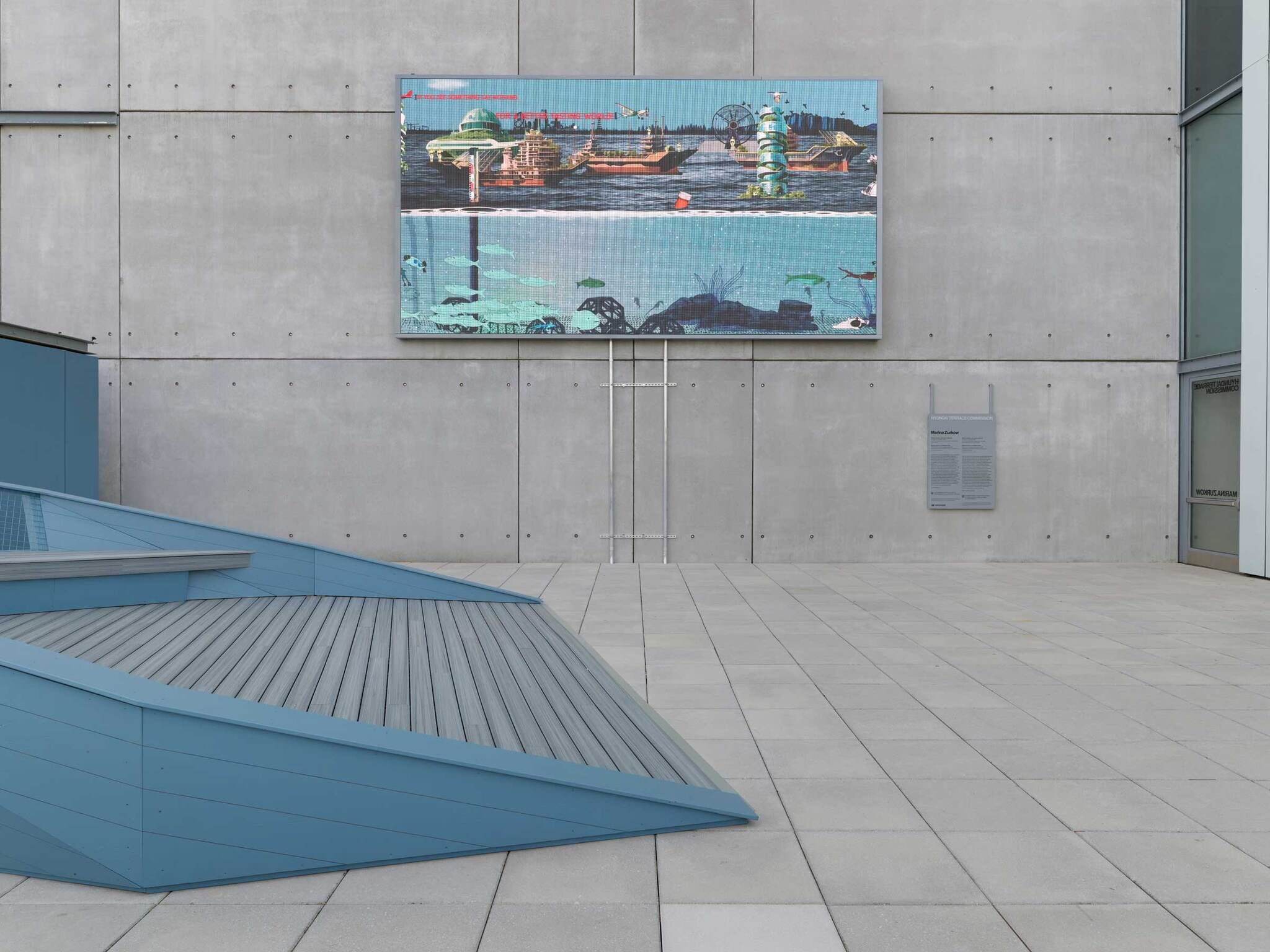 Large colorful mural showing a cityscape and underwater scene on a concrete wall in a modern outdoor plaza.