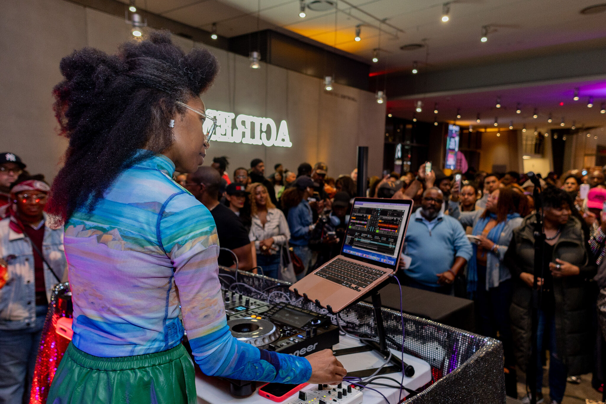 A DJ plays music on a laptop and mixer while a large crowd watches and takes photos.