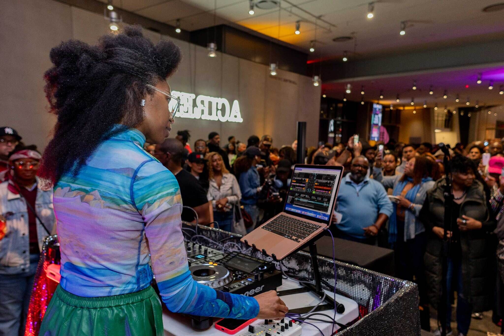 A DJ plays music on a laptop and mixer while a large crowd watches and takes photos at an indoor event.