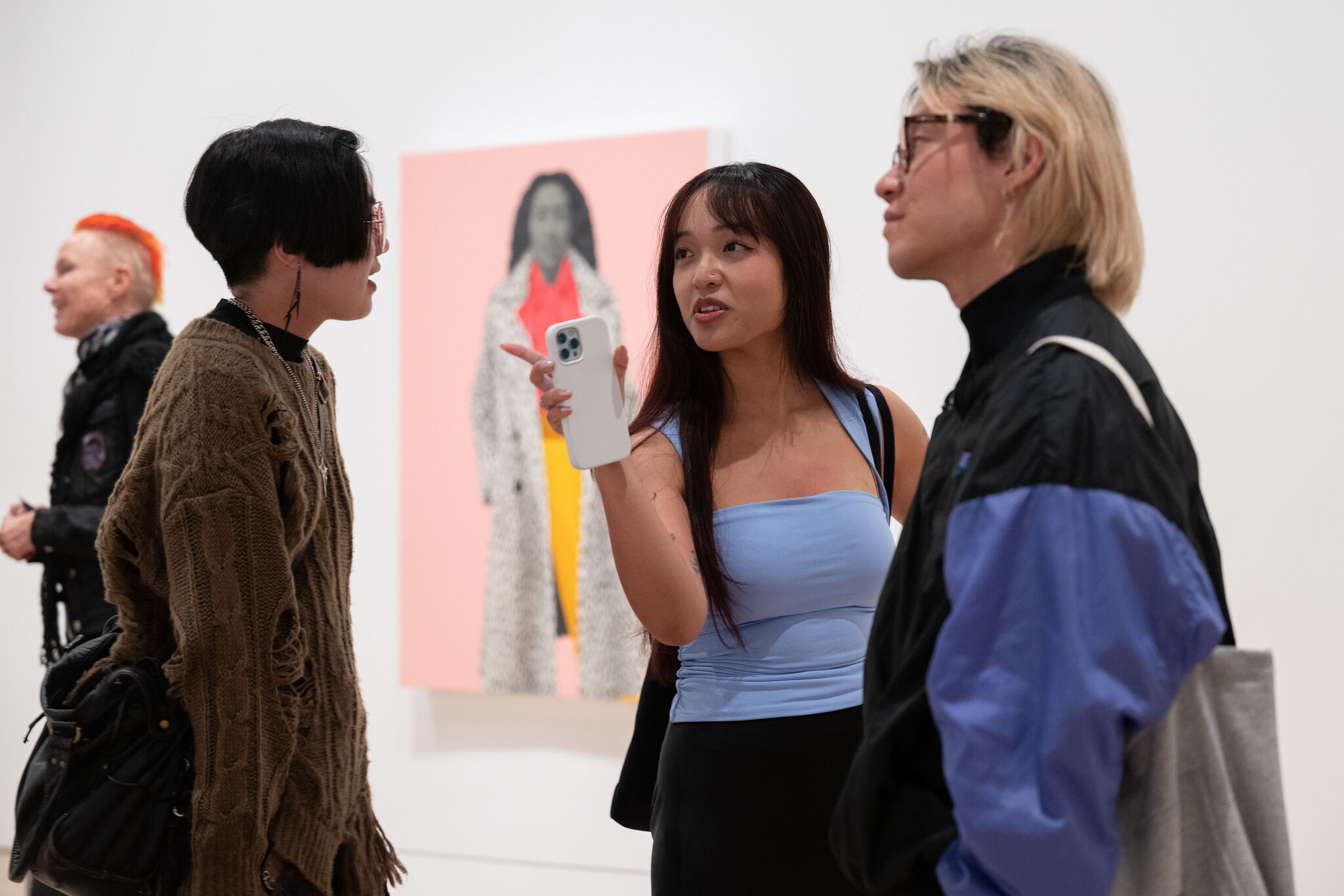Three people stand talking in an art gallery, with one person holding up a phone and gesturing.