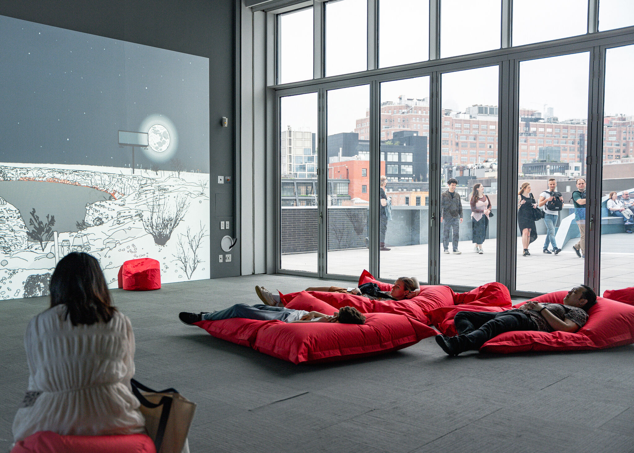 People relax on large red bean bags indoors, watching a wall projection of a moonlit landscape.