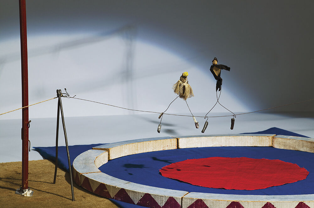 Two small puppets dressed as acrobats balance on a tightrope above a miniature circus ring.