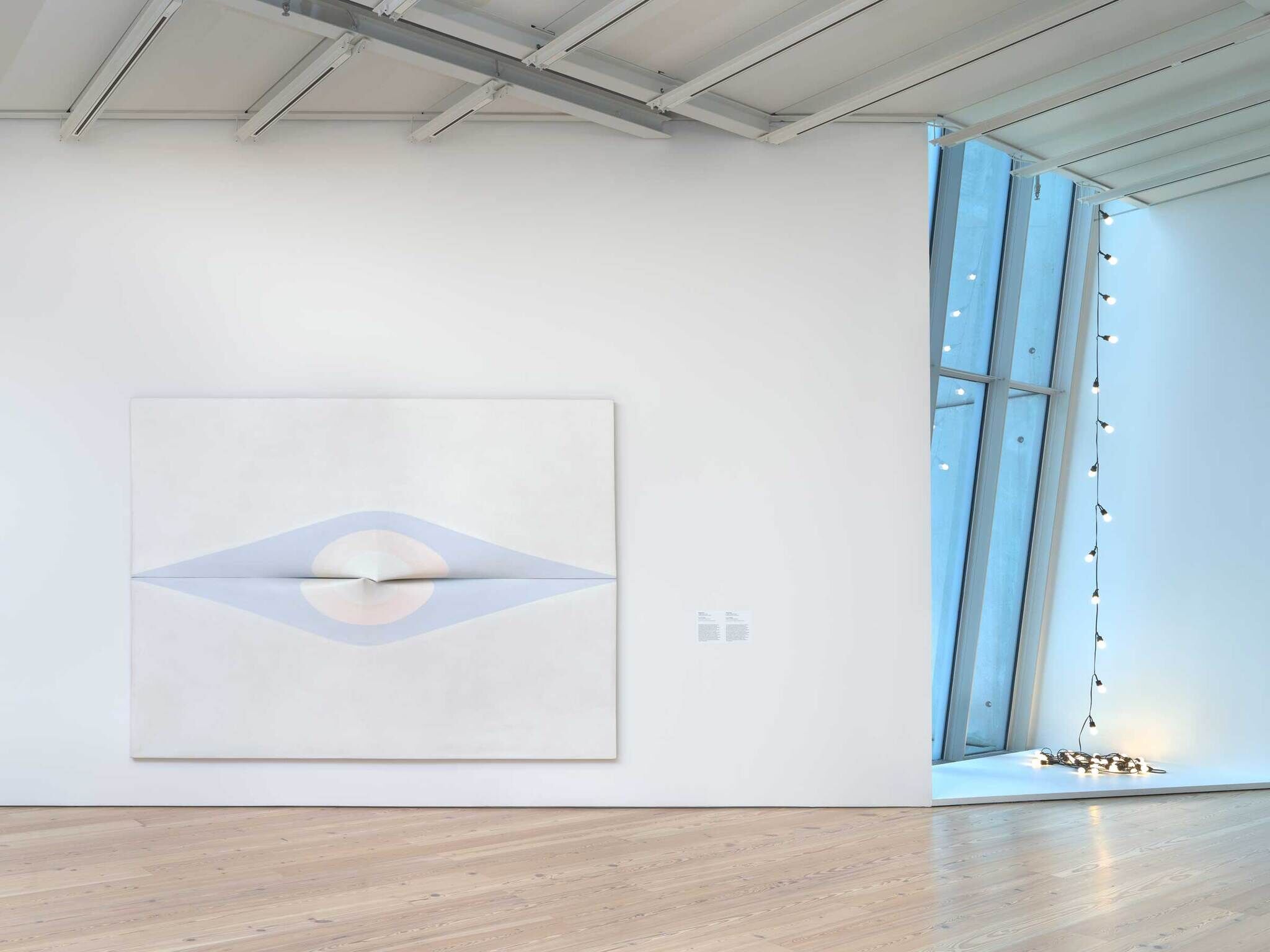 Large abstract painting with soft colors hangs on a white wall; string lights are placed near a window.