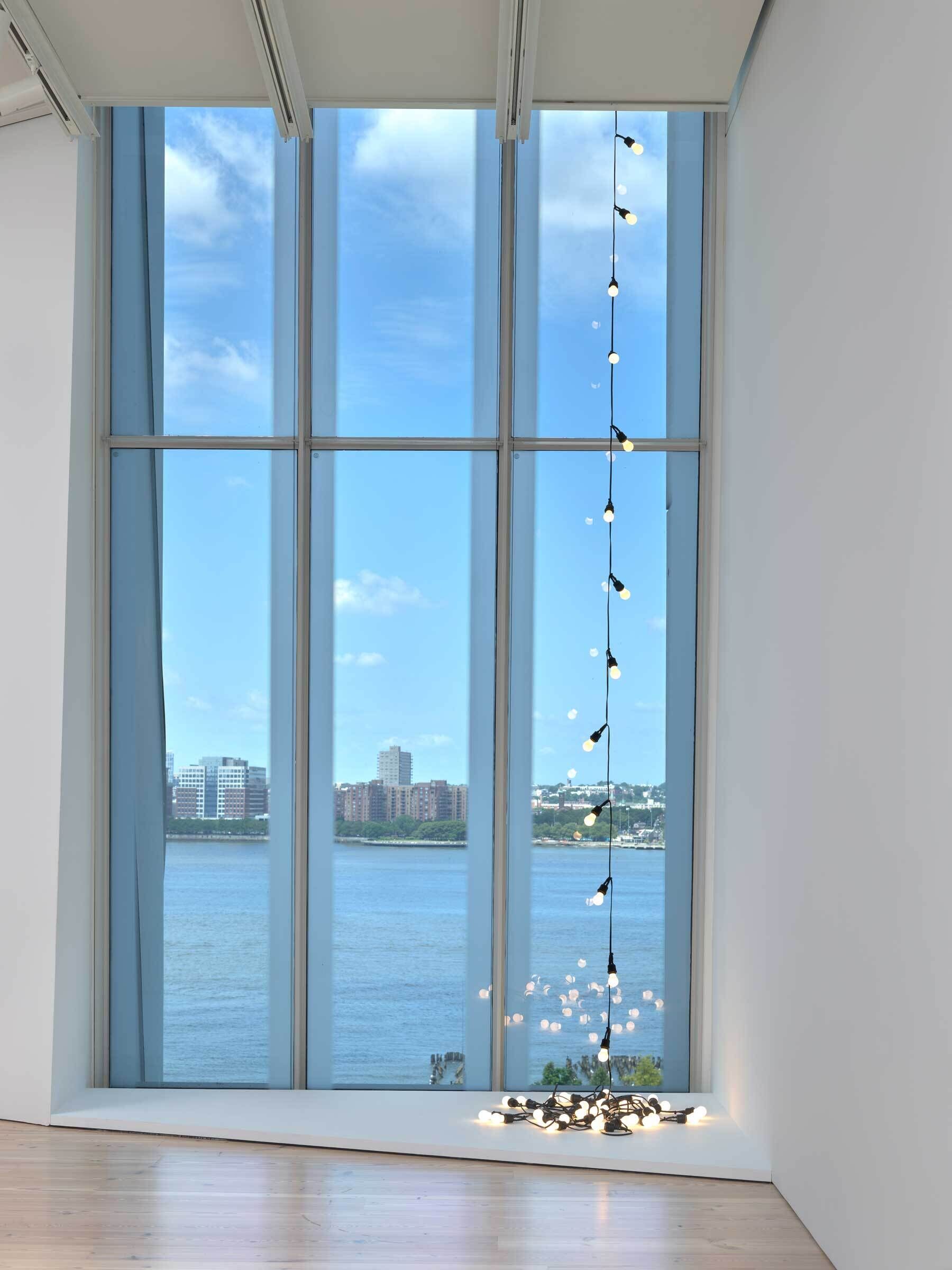 A string of lit lightbulbs draped on the right side of a window overlooking water and a clear blue sky.