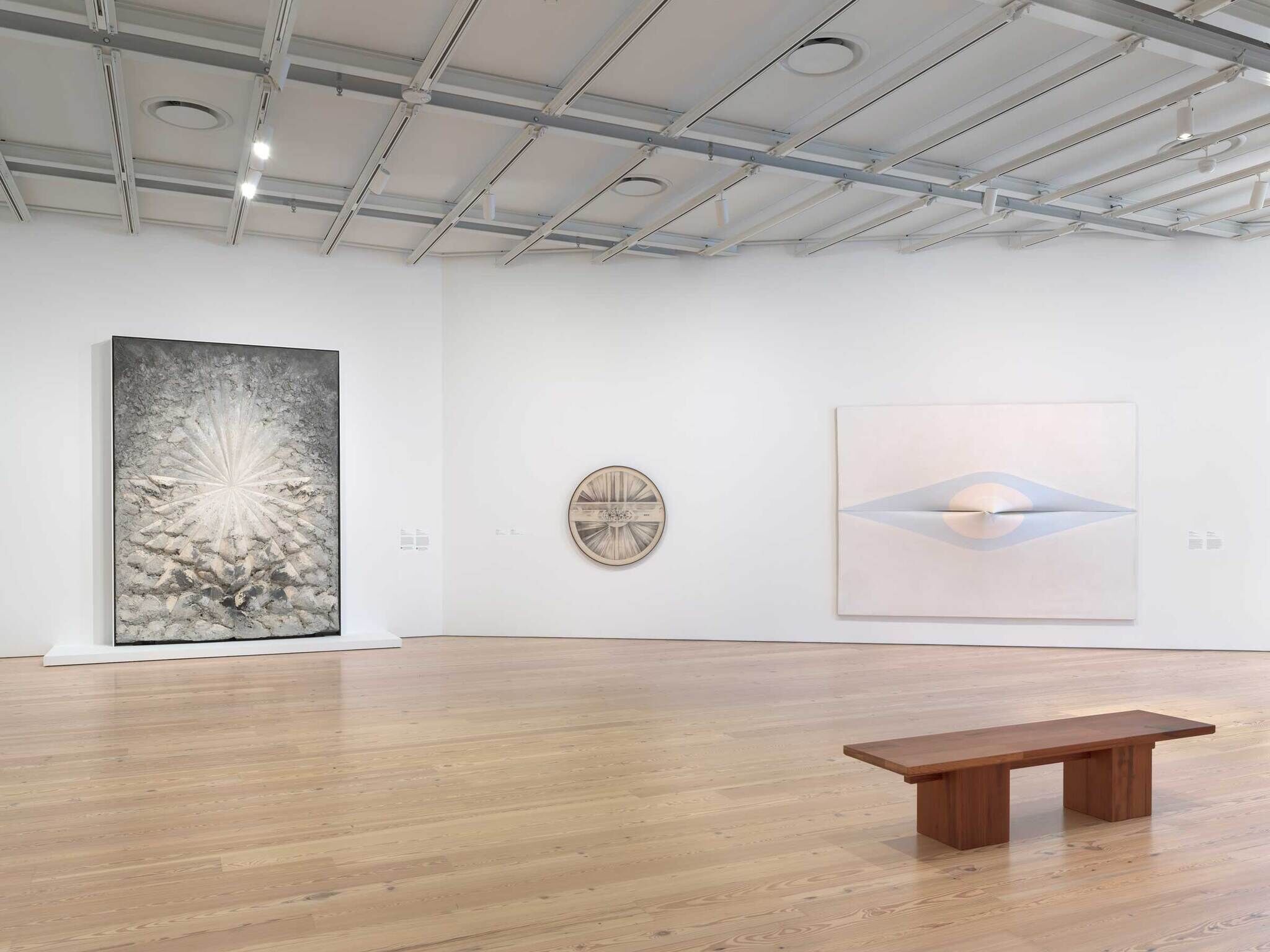Three abstract artworks hang on a white gallery wall with a wooden bench placed on the floor in front.